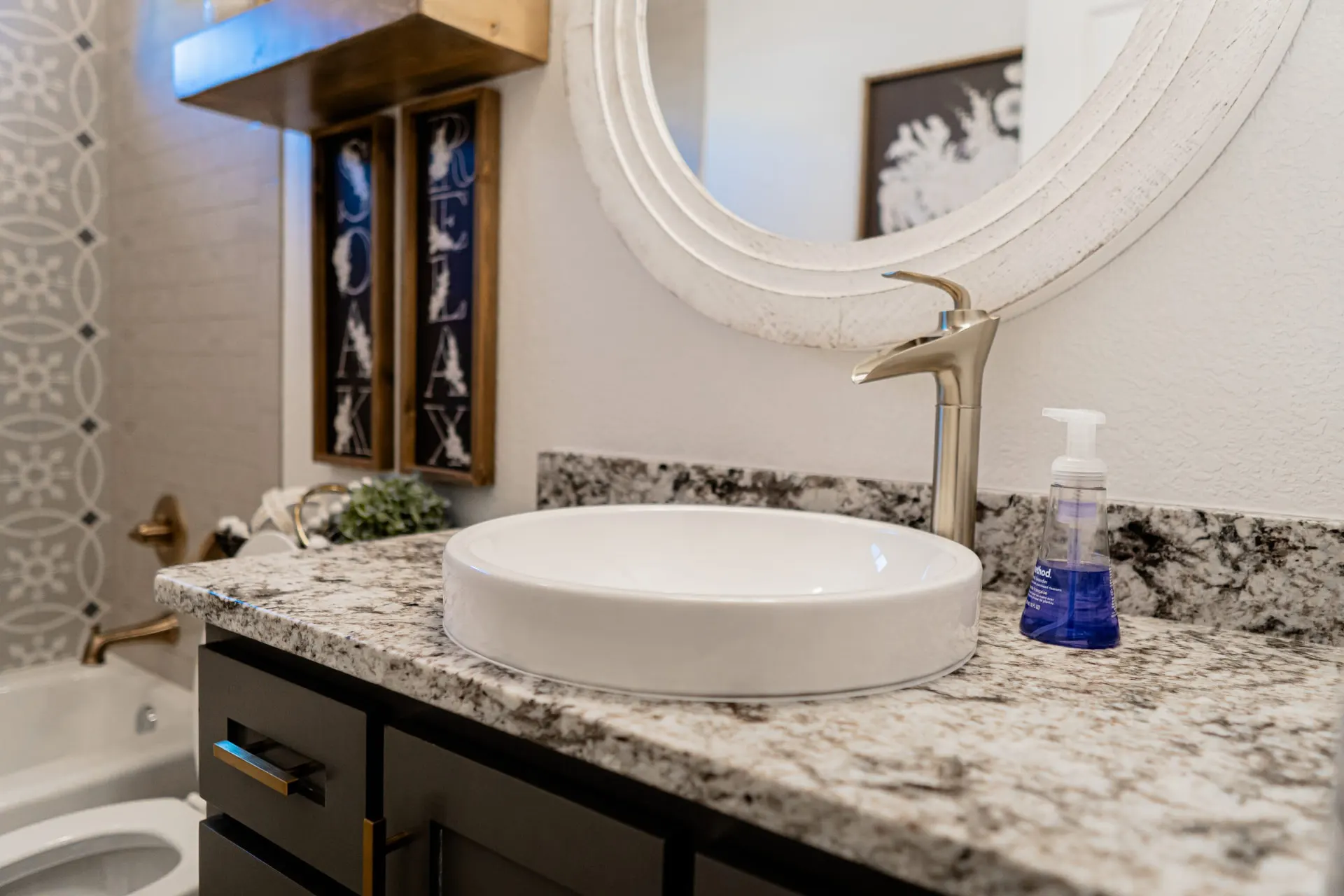 Bathroom vanity at the K Ave Farmhouse with a charcoal cabinet, speckled granite top, white round vessel sink, a distressed white round mirror and framed "Soak" and "Relax" signs