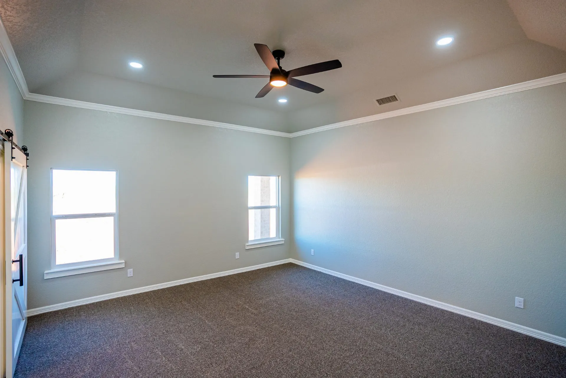 Empty bedroom at the Eliam St Home with a tray ceiling, dark ceiling fan, twin windows and fresh grey carpet