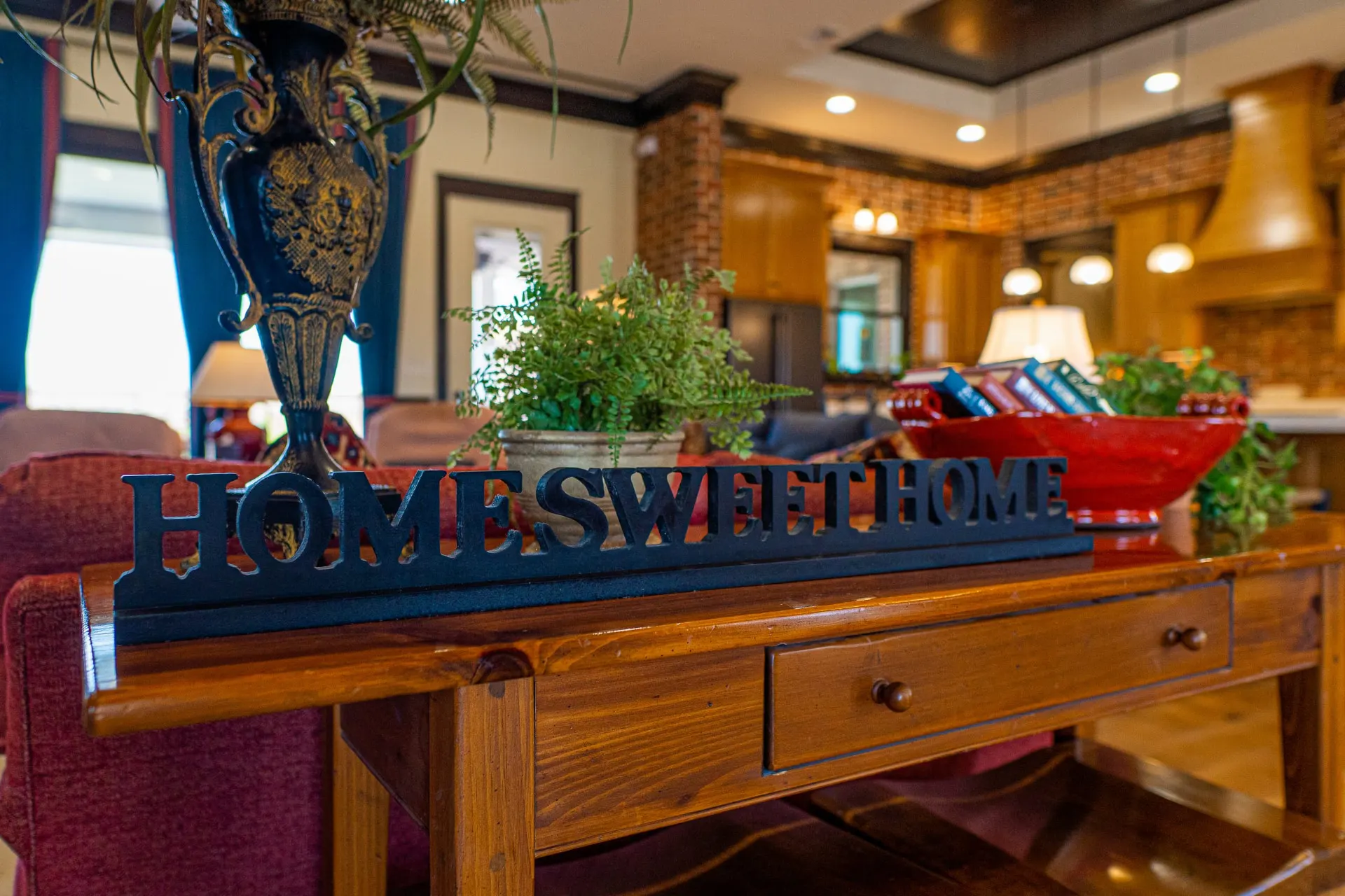 Console table behind the sofa at the Wraparound Porch Home with a "Home Sweet Home" cutout sign, a potted fern, a classical urn and a glossy red bowl