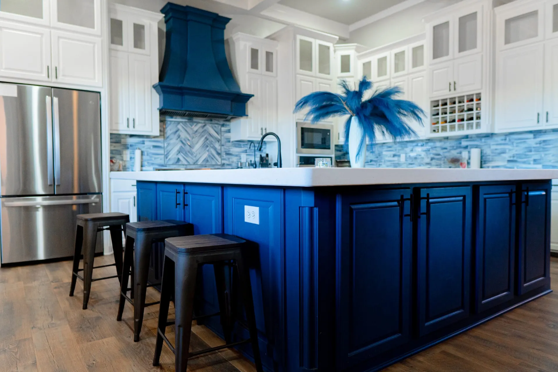 Kitchen at 8821 Shawn Dr with a navy island and black stools under a white quartz top, a navy range hood, white shaker cabinetry, a blue herringbone backsplash and a built-in wine rack