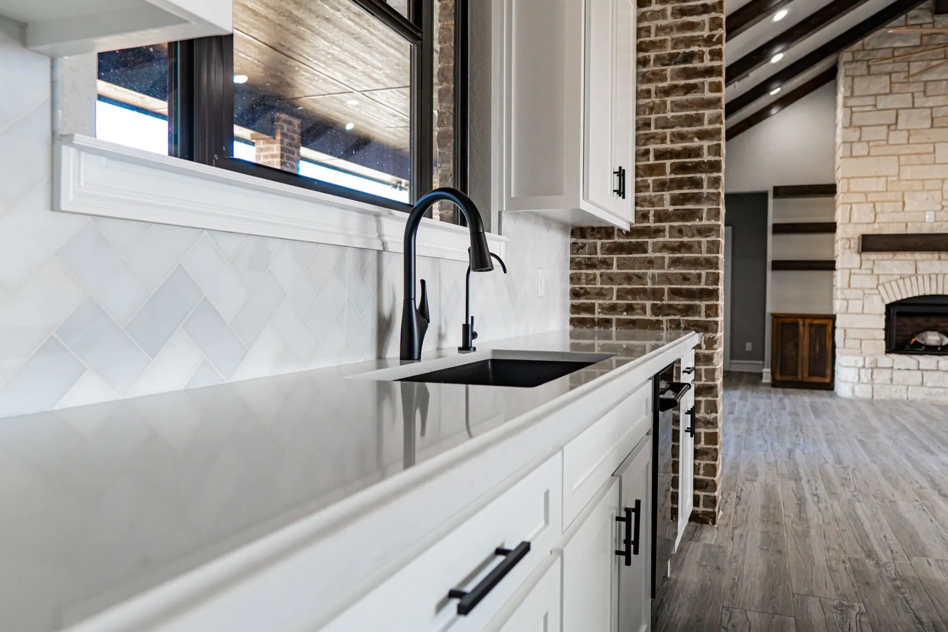 Sink run at the 12th St Home with a black faucet, white quartz and herringbone tile, the brick column and stone fireplace beyond
