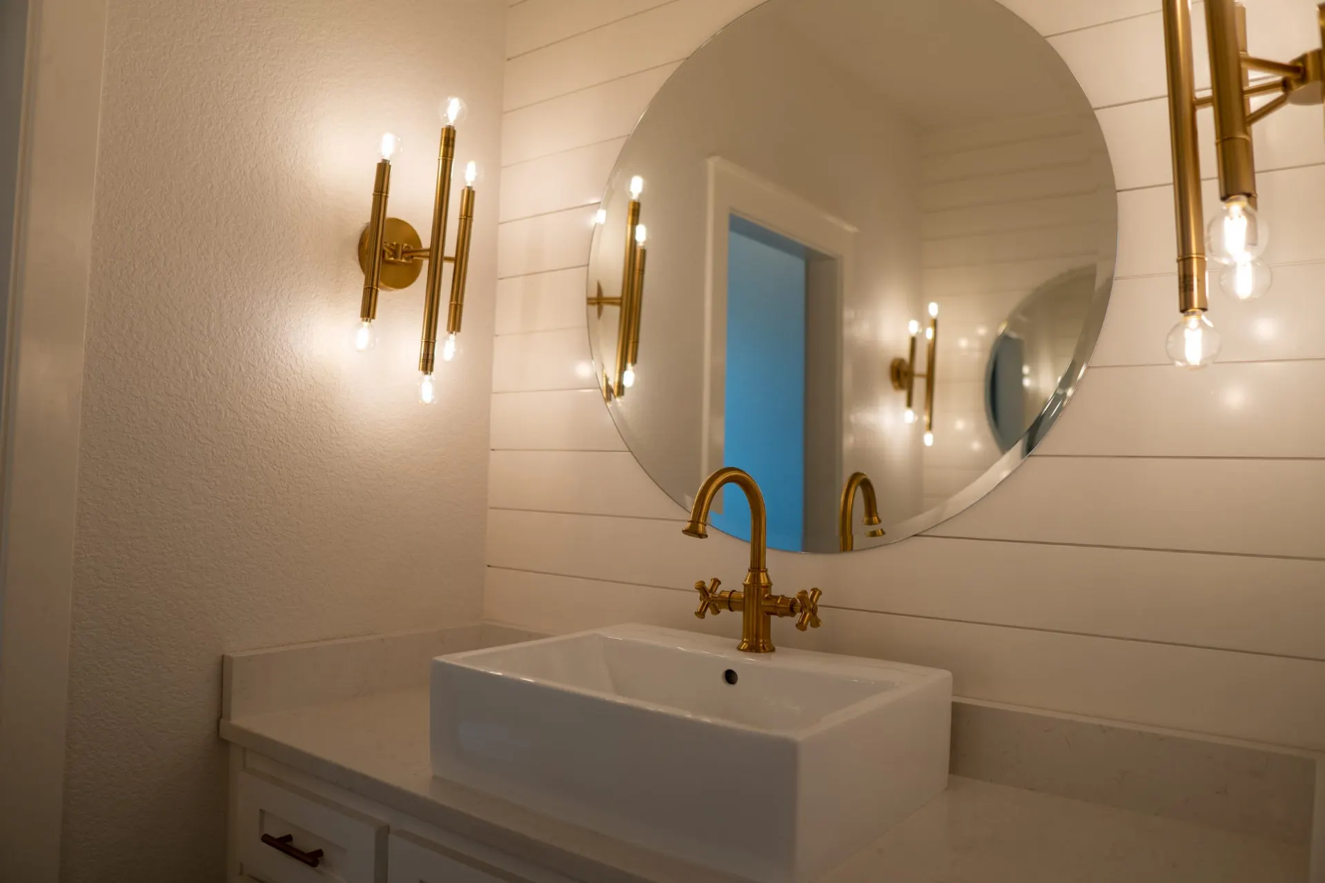 Bathroom vanity at Sarai Home with a white vessel sink under a brushed-gold cross-handle faucet and round mirror, lit by gold stem sconces on white shiplap