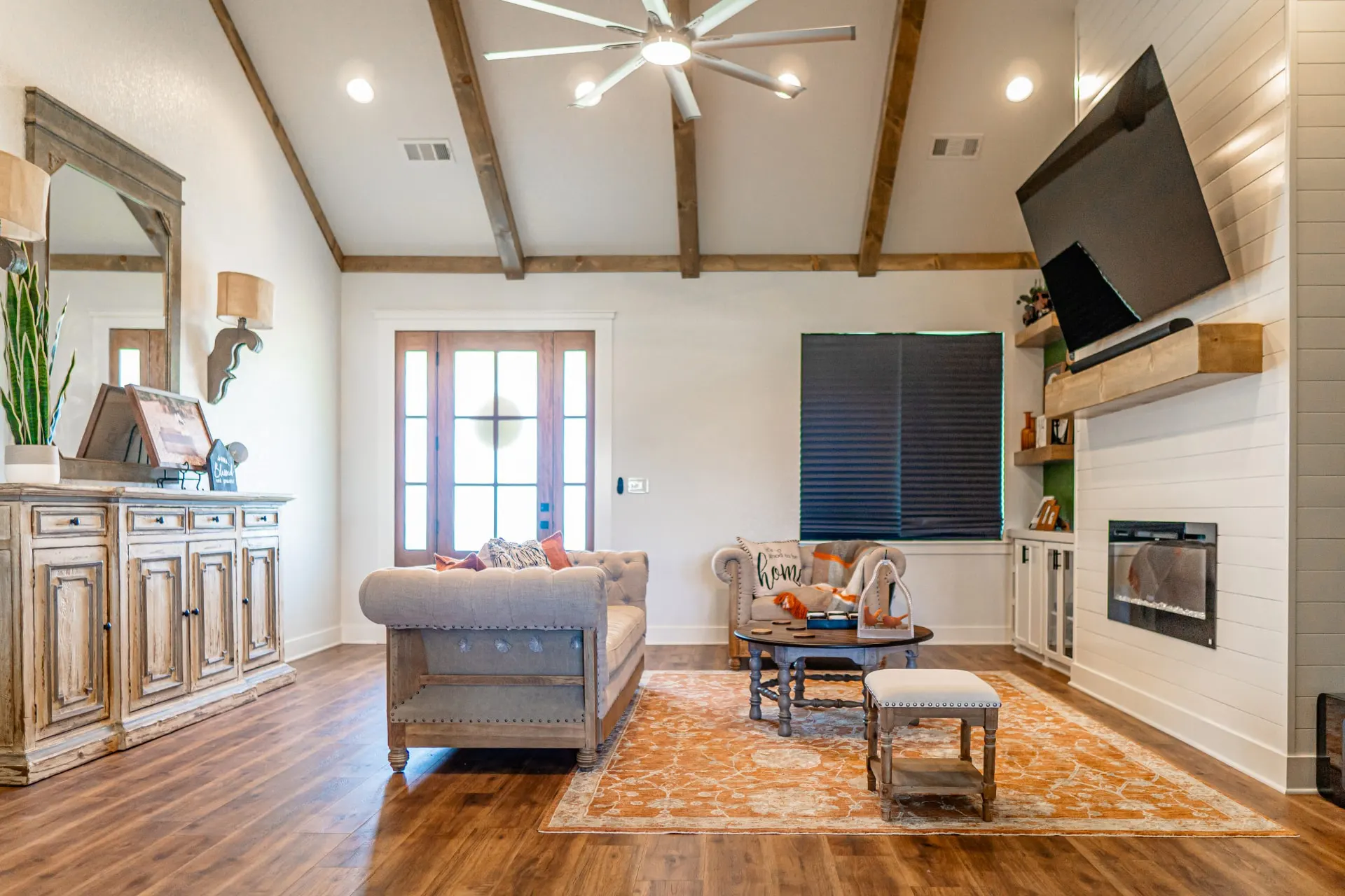 Living room at the K Ave Farmhouse from the entry, with wood ceiling beams, a fireplace under a mounted TV and a navy roller shade