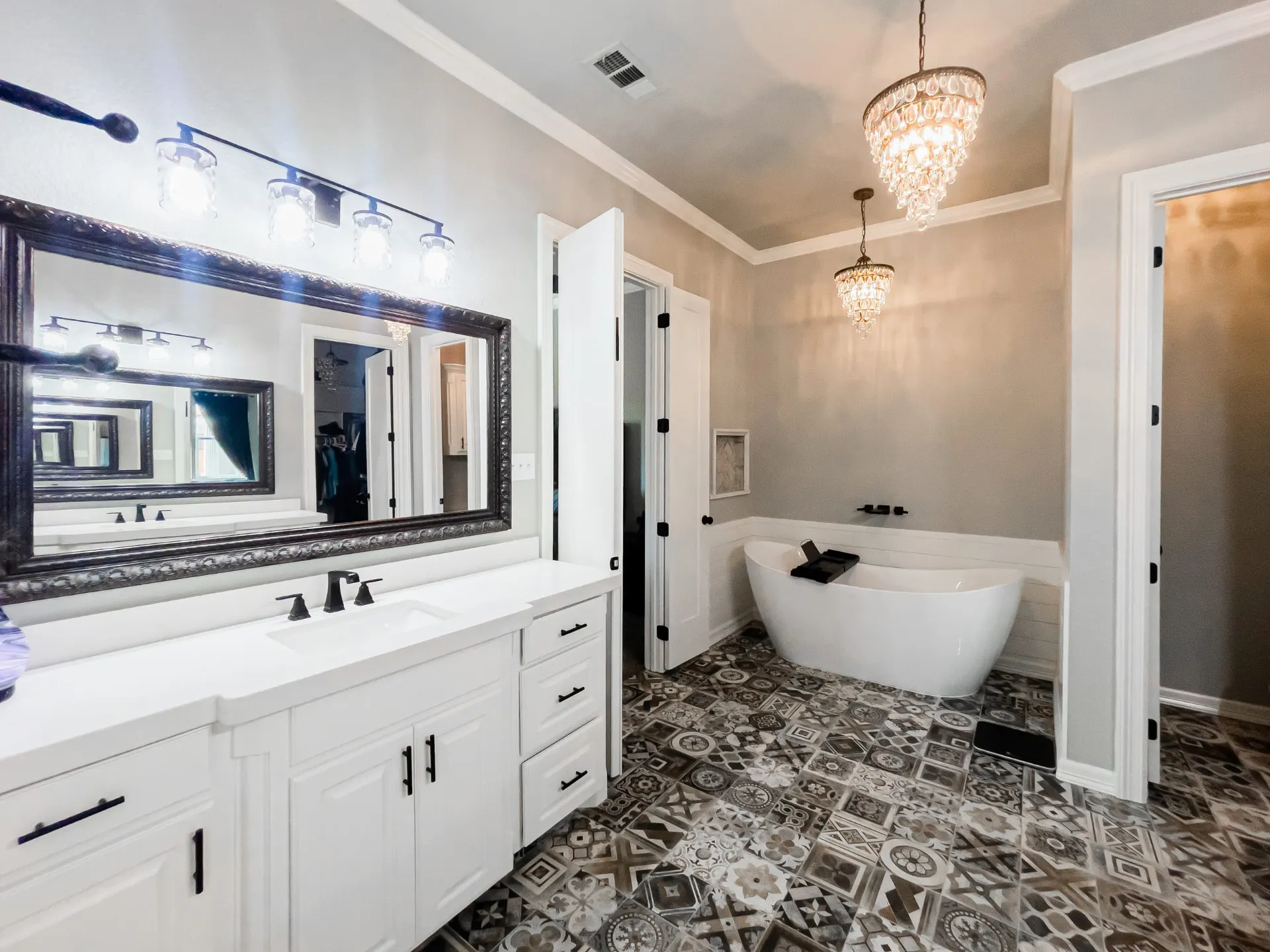 Primary bath at 8821 Shawn Dr with a long white vanity under a four-light bar, a freestanding tub beneath two crystal chandeliers and grey-and-cream patterned floor tile