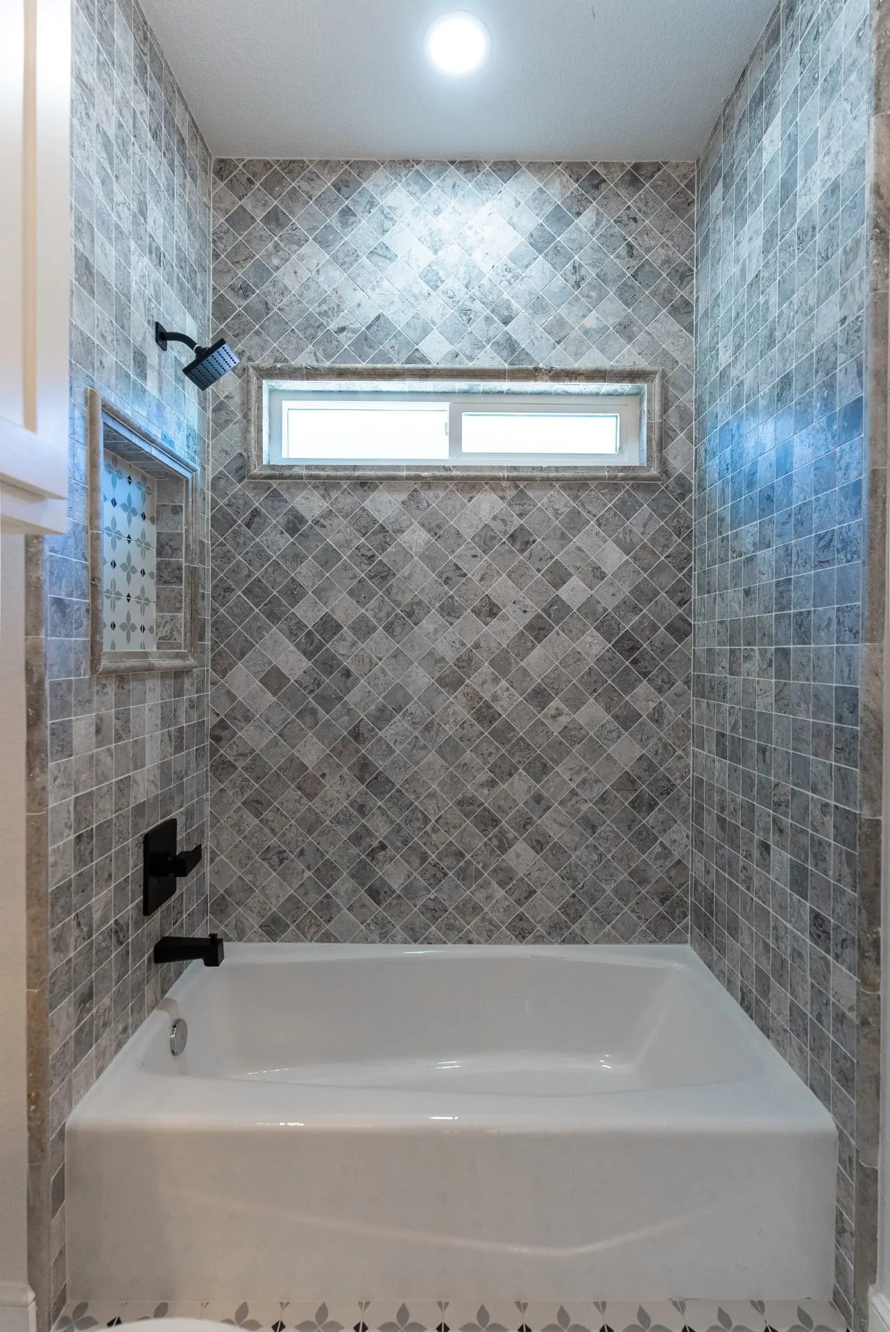 Tub and shower at 718 K Ave framed in diamond-set gray stone tile with a transom window and patterned niche