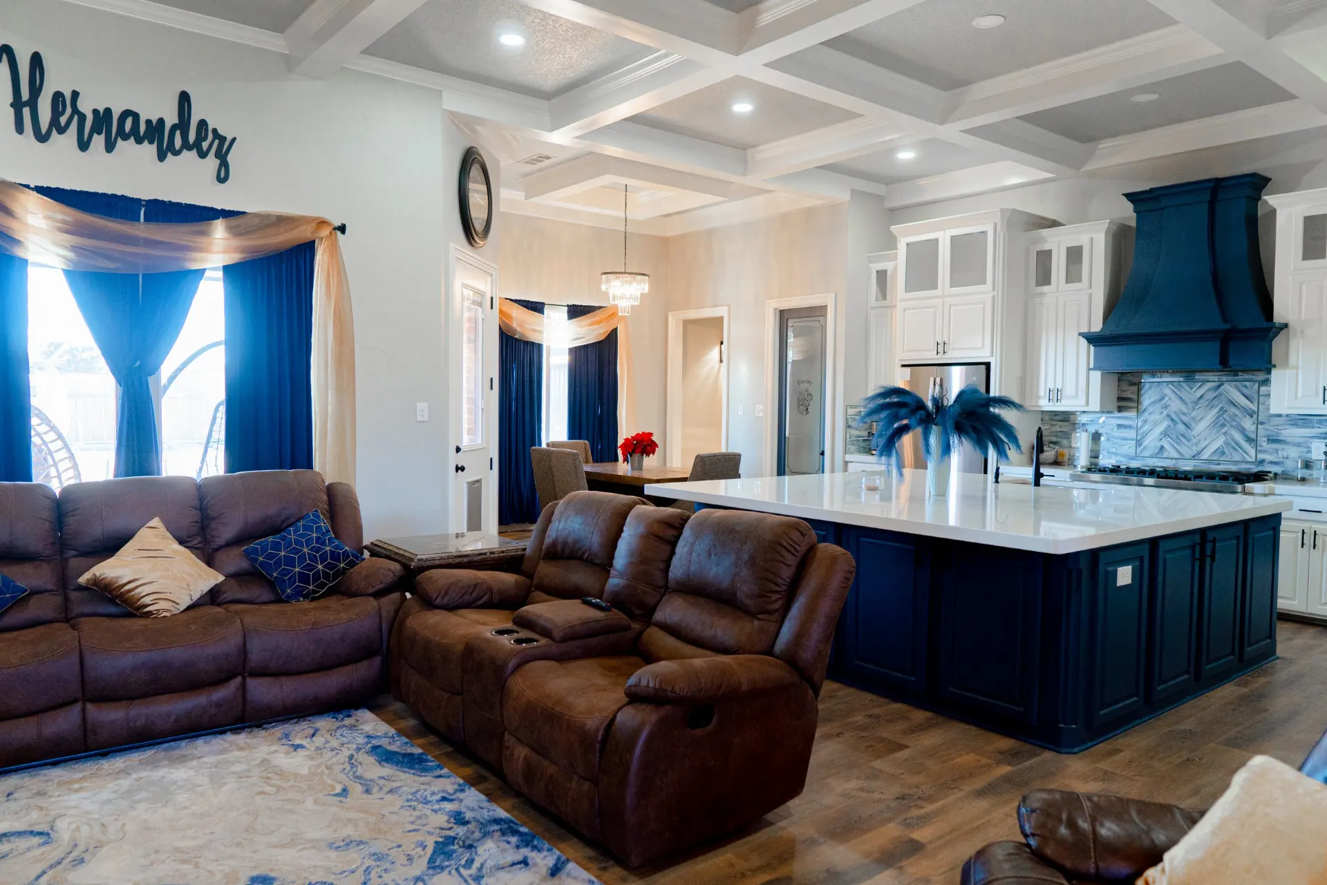 Open living room and kitchen at 8821 Shawn Dr under a coffered ceiling, with a large navy island, a navy hood and a dining area with a crystal chandelier beyond