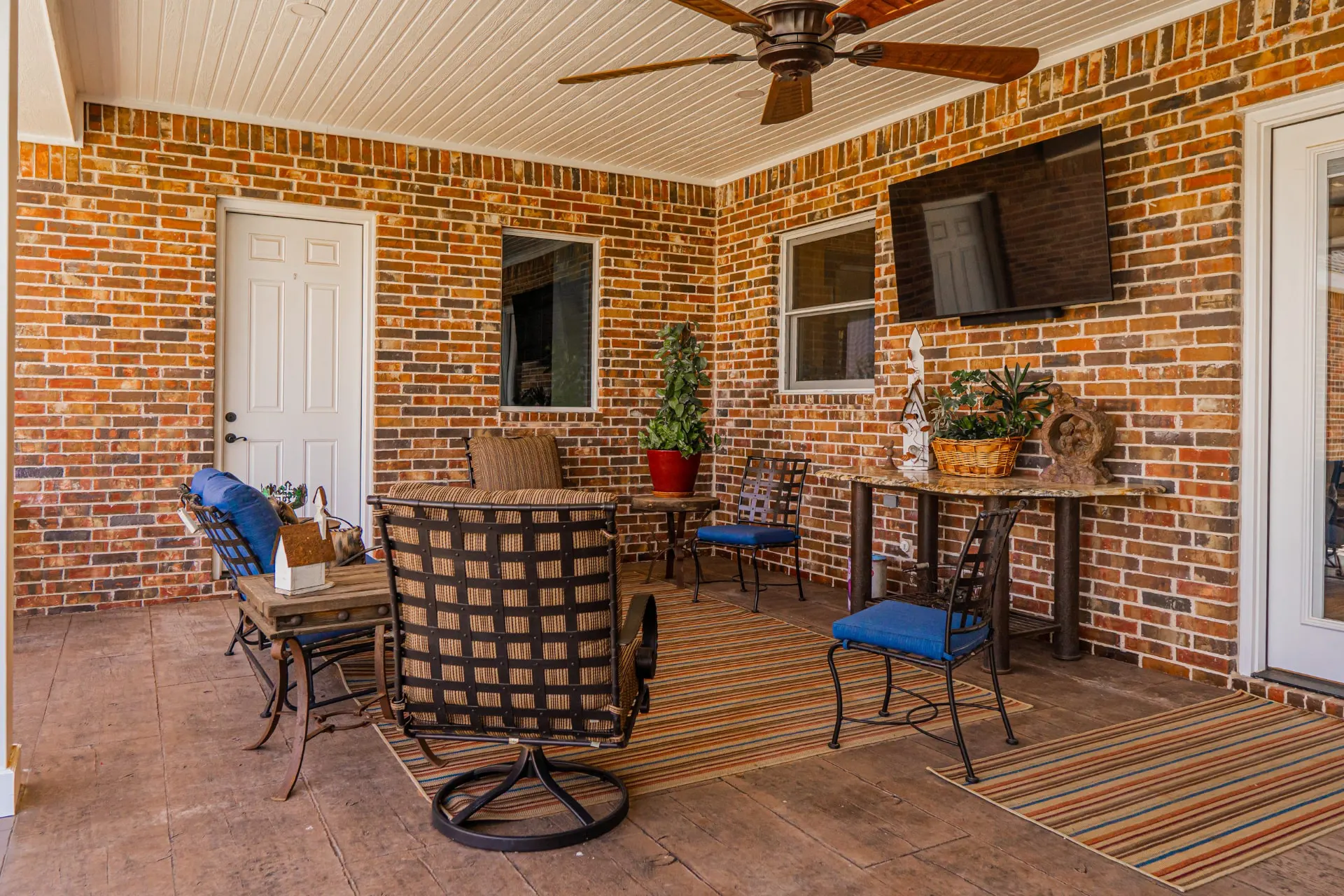 Covered patio seating area at the Wraparound Porch Home with a wall-mounted television, iron chairs and striped rugs on stamped concrete