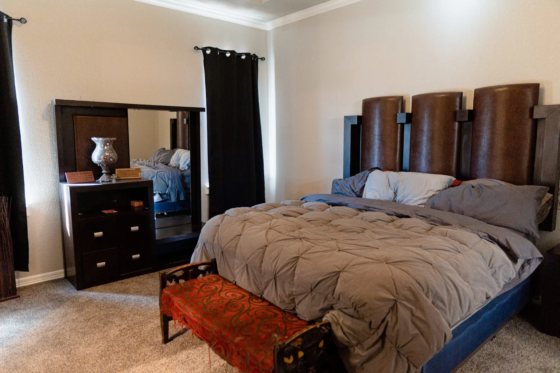 Primary bedroom at 8821 Shawn Dr with a tall dark-framed headboard, grey bedding, a red patterned bench at the foot of the bed and a dark wood dresser with an integrated mirror