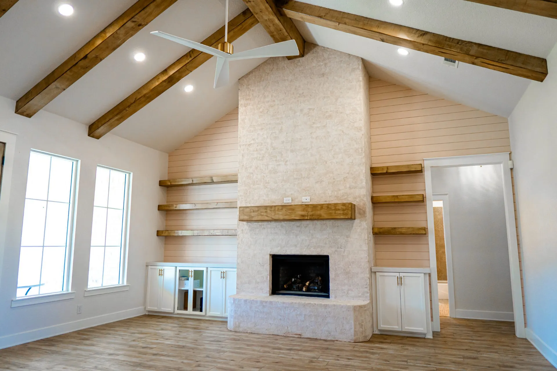 Vaulted great room at Sarai Home with wood ceiling beams and a full-height plastered fireplace under a rough-hewn wood mantel, flanked by shiplap walls and floating shelves