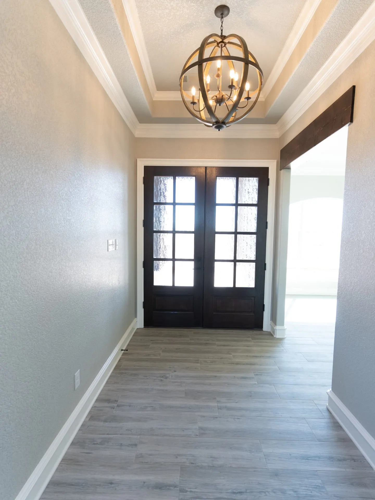 Entry foyer of the Eliam St Home with dark double doors, a beaded wood globe chandelier in a coffered tray ceiling and grey wood-look tile