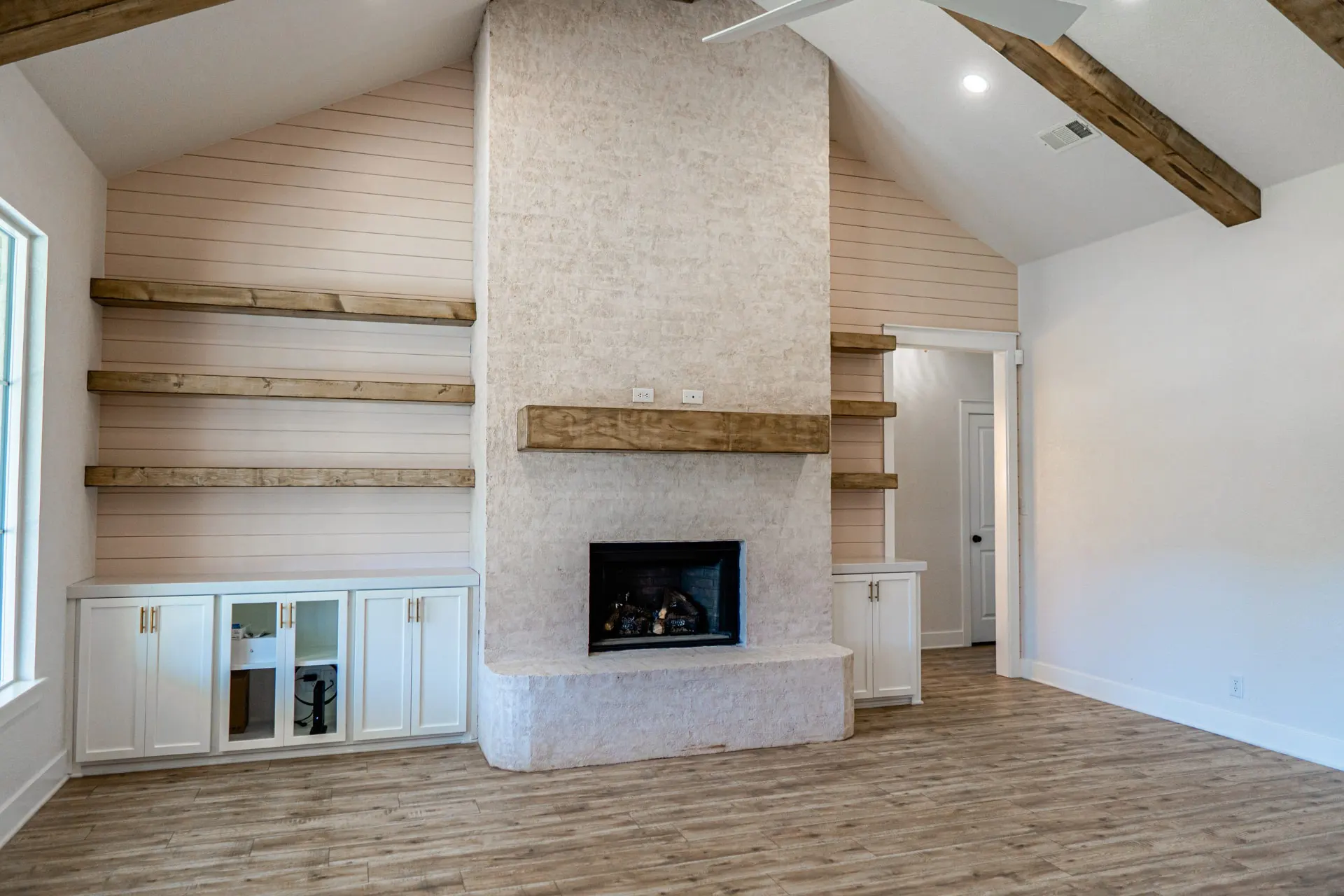 Fireplace wall at Sarai Home with white built-in cabinets and glass doors on either side of the plastered fireplace and floating wood shelves