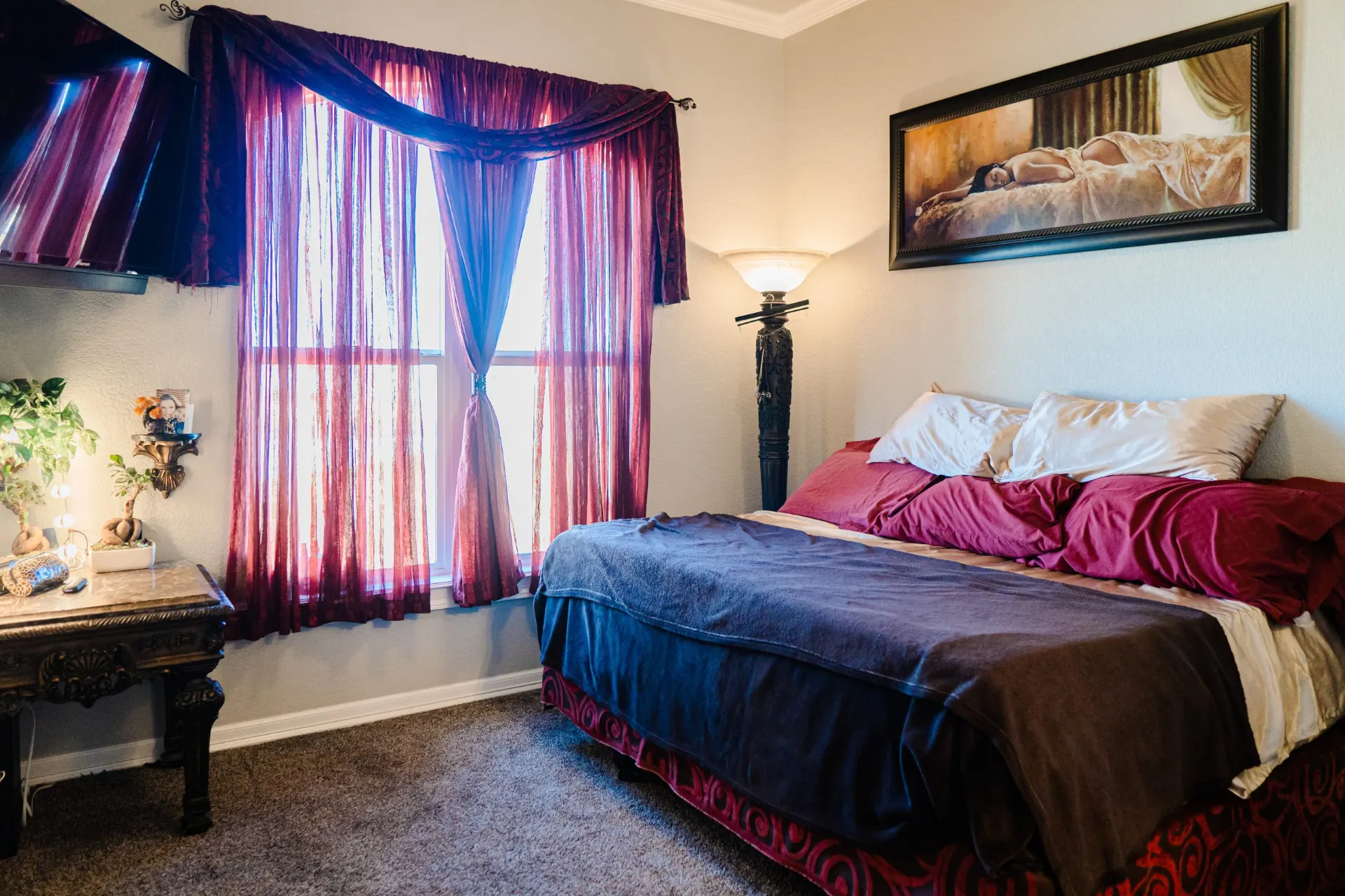 Bedroom at 8821 Shawn Dr with deep red bedding and sheer burgundy drapery, a carved dark wood bedside table and a corner-mounted TV