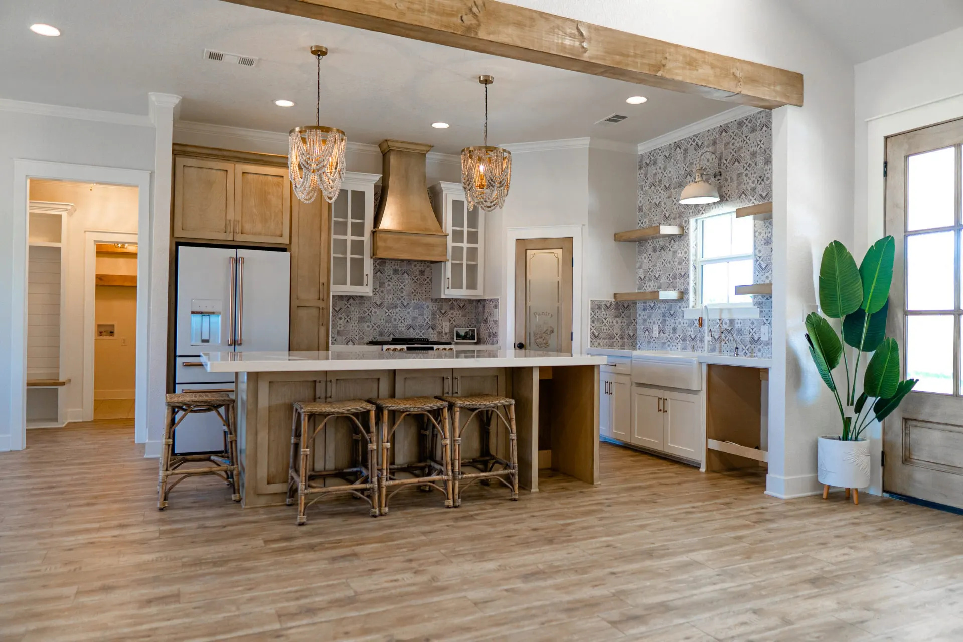 Open kitchen at Sarai Home with a large wood island under a white quartz top, rattan counter stools and two beaded chandeliers, with a patterned-tile backsplash wall and farmhouse sink beyond