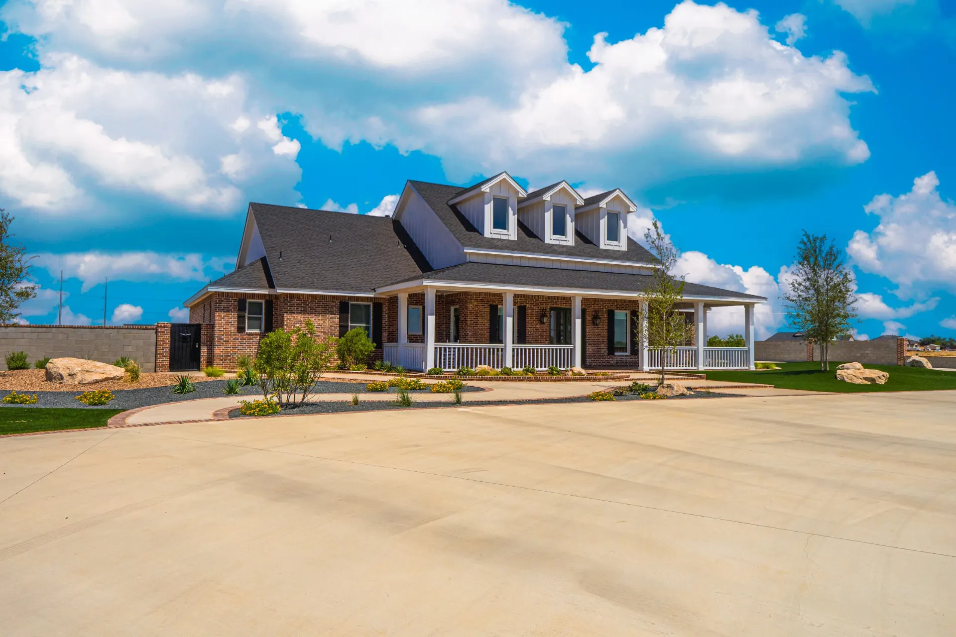 Angled front exterior of the Wraparound Porch Home in red brick with white dormers, a covered front porch and a curved landscaped drive under blue skies
