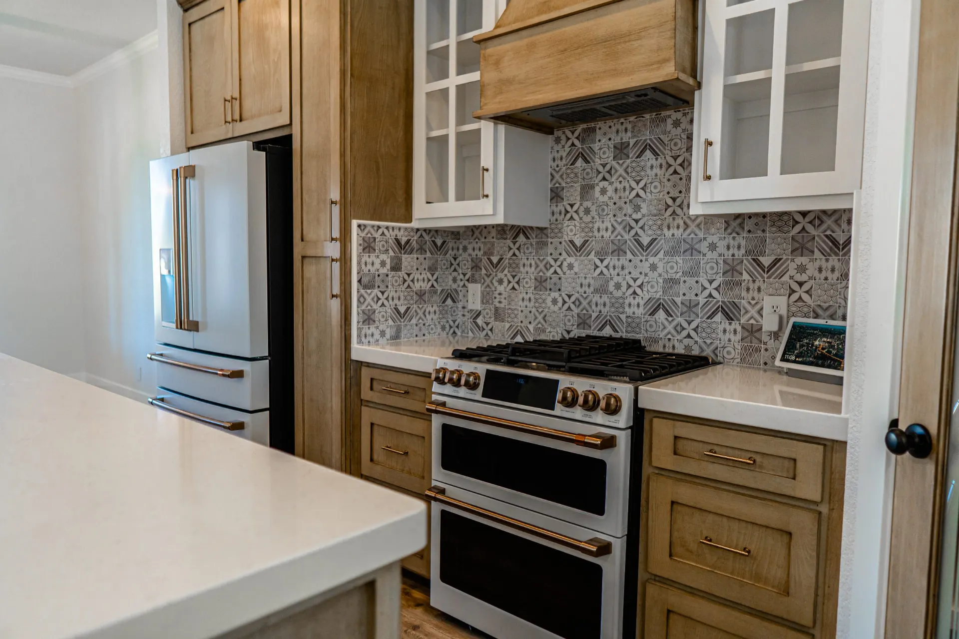 Cooking zone at Sarai Home with a stainless gas range under a wood range hood, white glass-front uppers and a patterned-tile backsplash