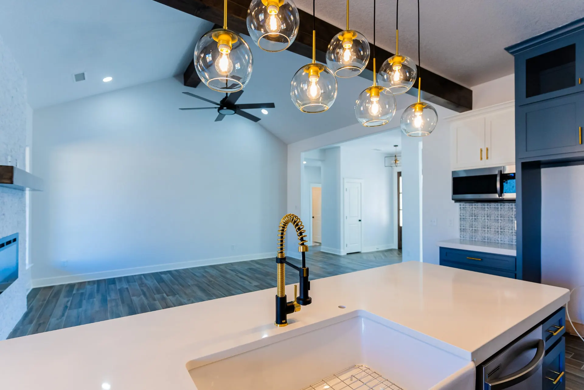 Kitchen island at 718 K Ave with a white countertop, undermount sink and black-and-brass coil faucet, under a cluster of glass globe pendants hung from a rough-hewn beam, with slate blue and white cabinetry and the open living room beyond