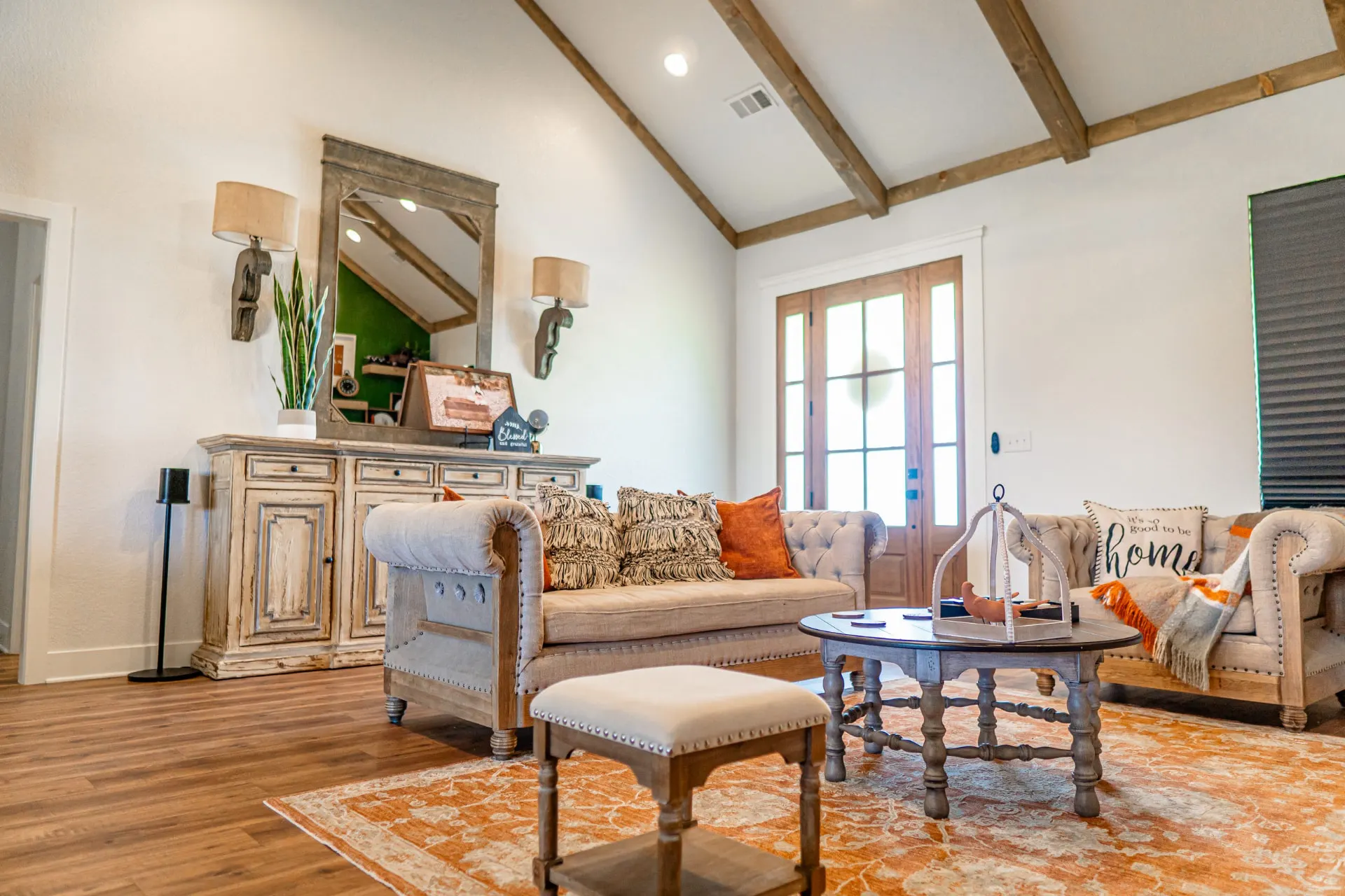 Living room at the K Ave Farmhouse under a beamed vaulted ceiling with an antique sideboard, wooden front doors and orange-accent pillows