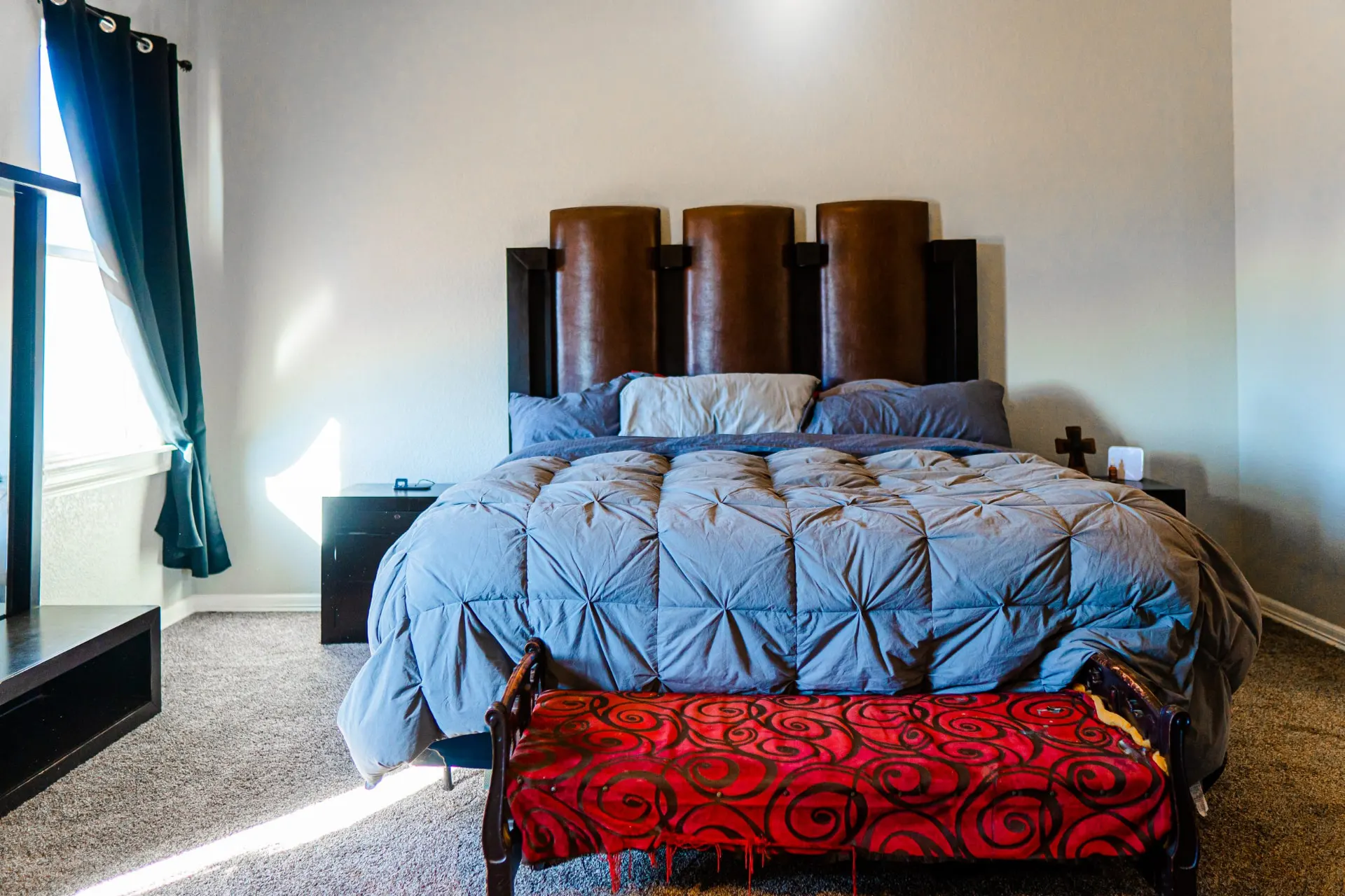 Primary bedroom at 8821 Shawn Dr facing a three-panel leather headboard with grey bedding and an ornate red-and-black bench, flanked by dark nightstands