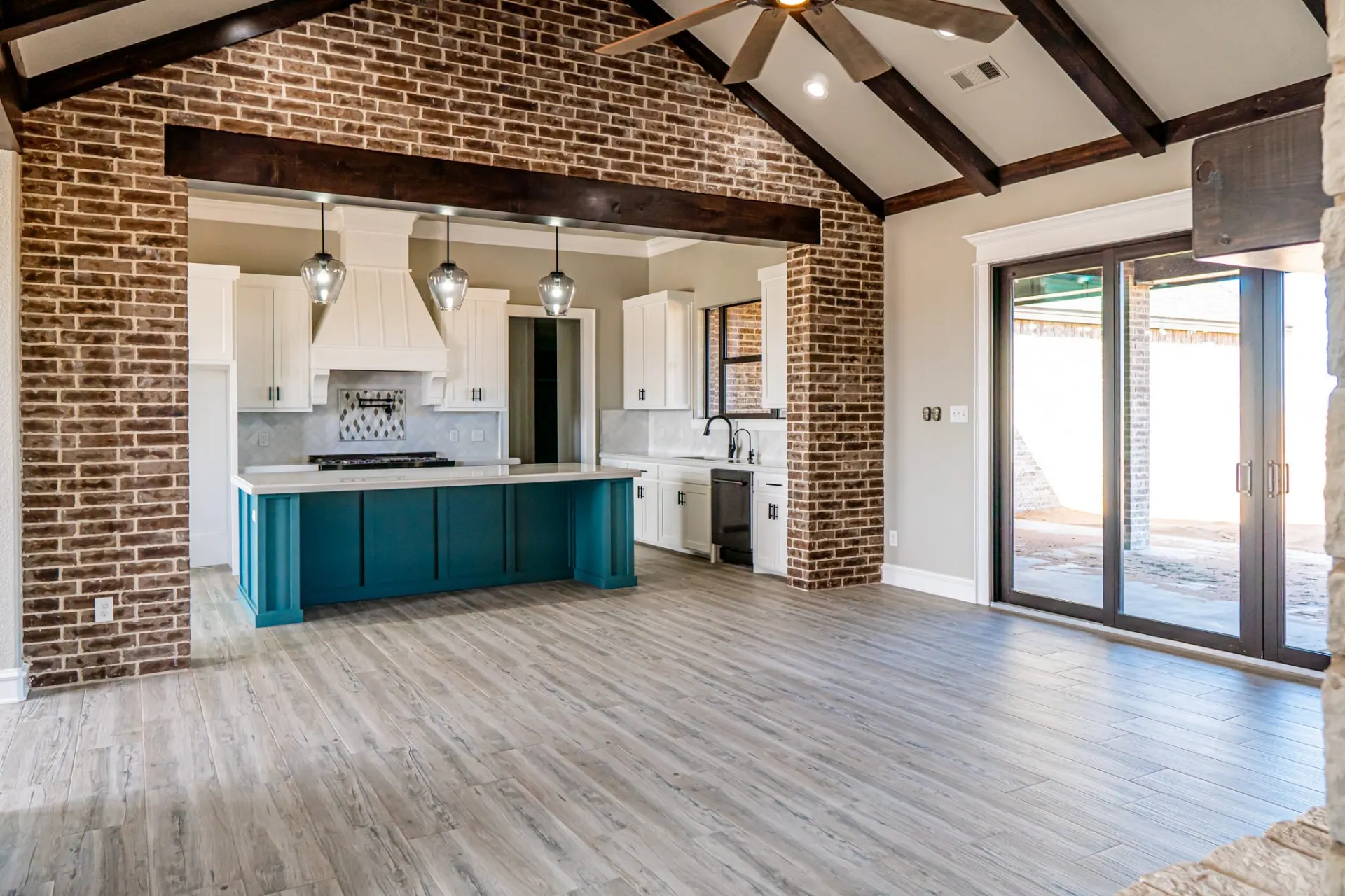 Open kitchen and living room at the 12th St Home seen through a rustic brick archway with a heavy timber header, the teal island and white hood beyond