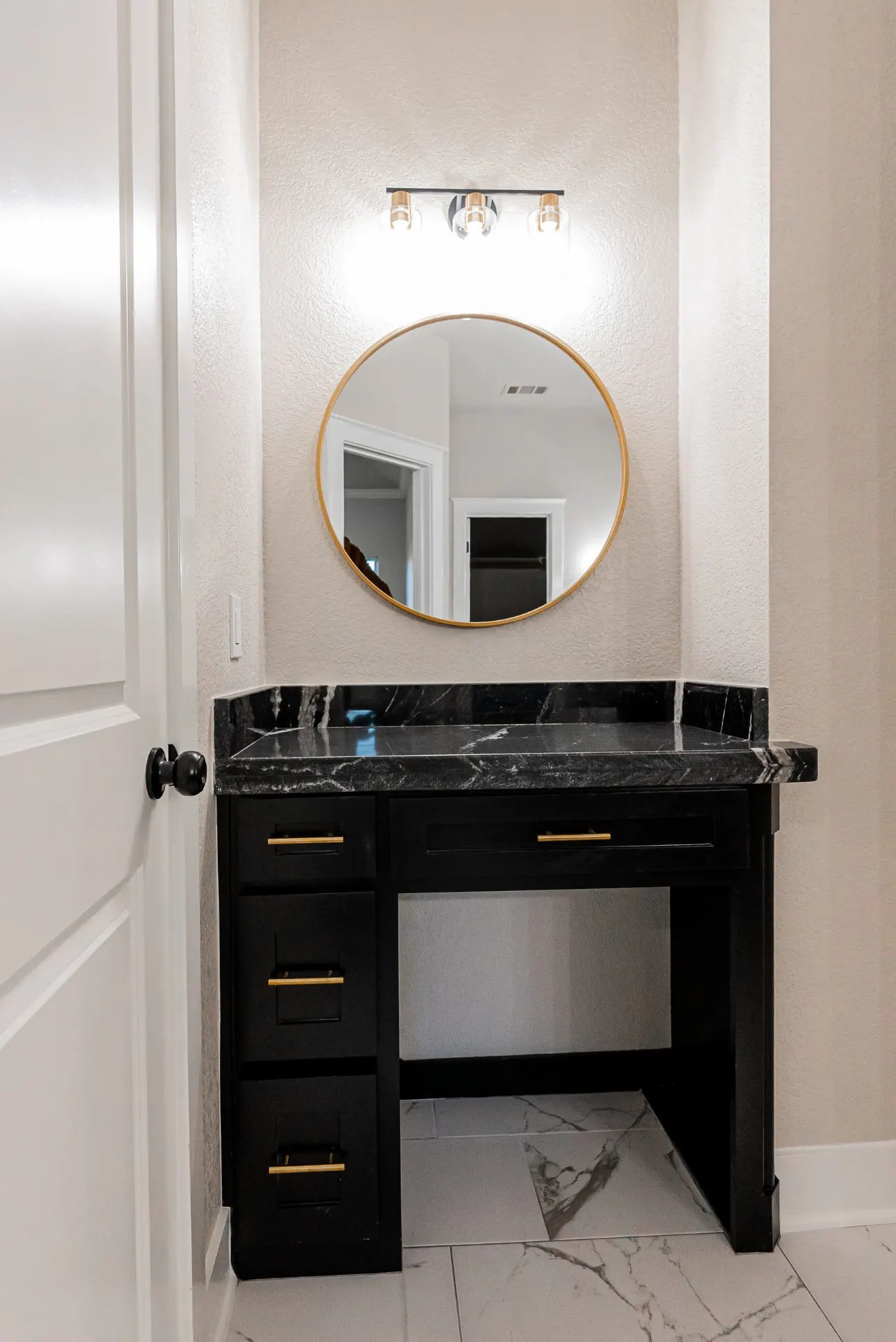 Bathroom vanity nook at 708 K Ave with black cabinetry, a black marble counter, gold-framed round mirror and gold sconces over marble-look floor tile