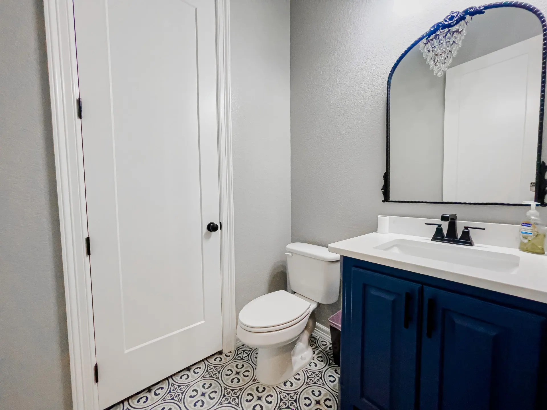 Powder room at 8821 Shawn Dr with a navy shaker vanity, matte black hardware, a black arched mirror and Mediterranean-style patterned floor tile