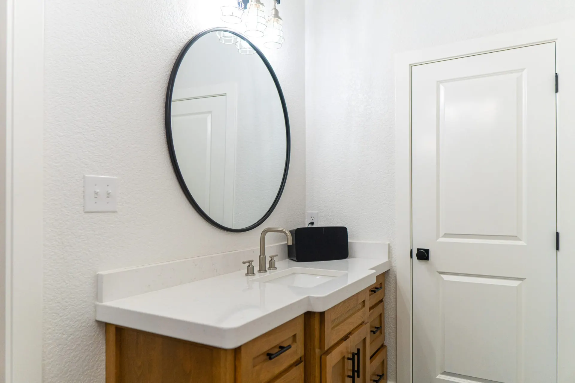 Vanity detail at the K Ave Farmhouse with a black oval mirror, brushed nickel faucet, black hardware and a wireless speaker on the quartz counter