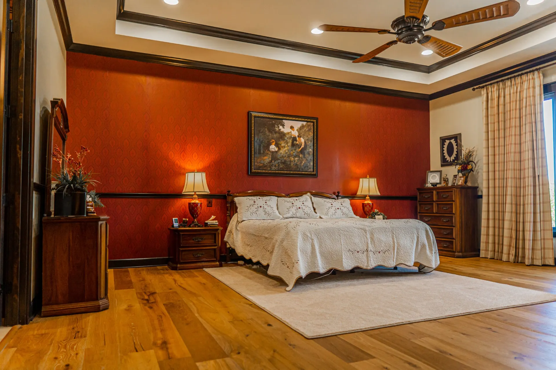 Primary bedroom at the Wraparound Porch Home with a red patterned accent wall, tray ceiling and wide-plank wood floors