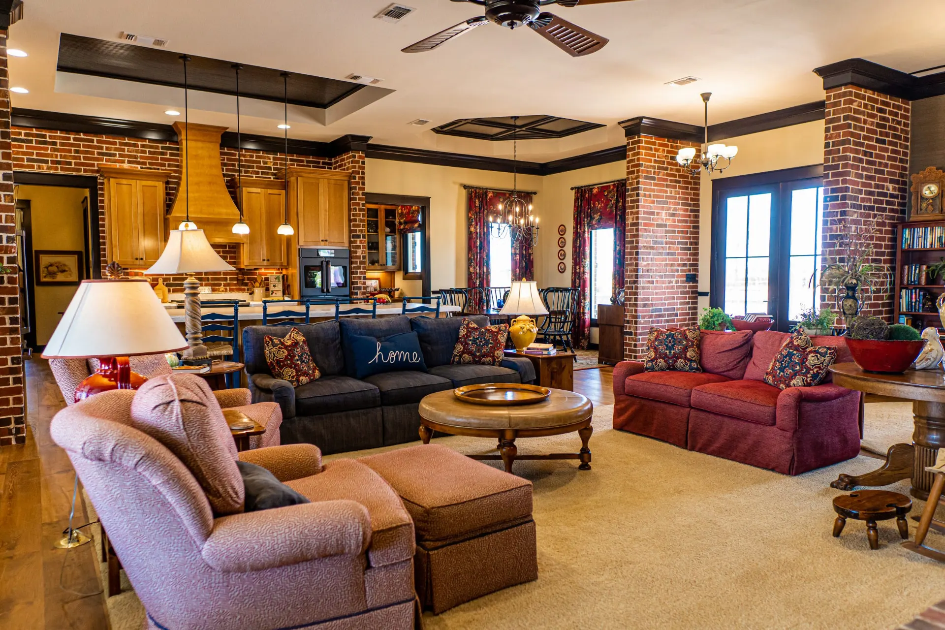 Wide living room view at the Wraparound Porch Home with brick pillars, a coffered dark wood tray ceiling, blue and red sofas and the kitchen beyond