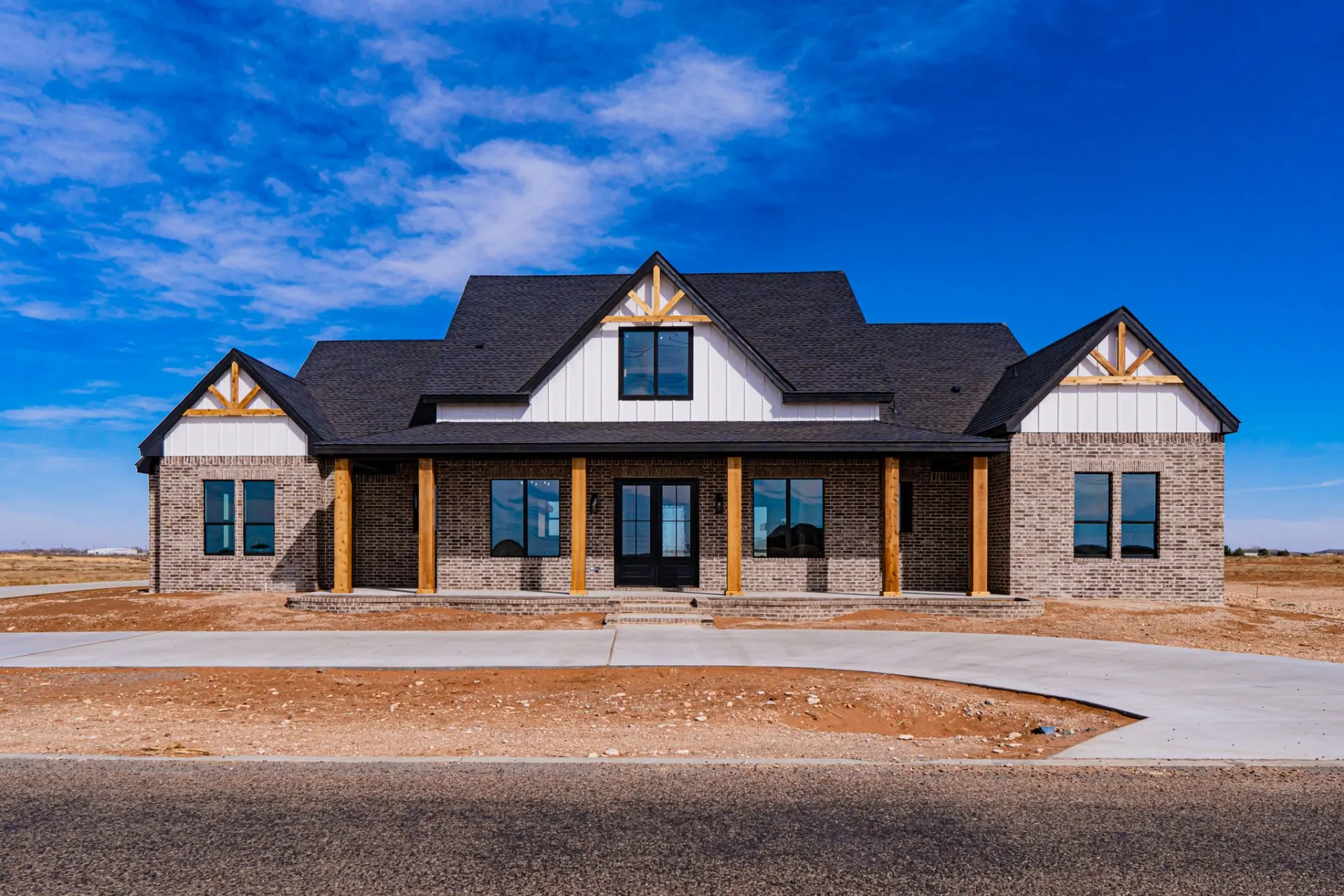 Front elevation of the 12th St Home with grey brick, white board-and-batten gables, a charcoal shingle roof and a deep covered porch on six natural wood columns