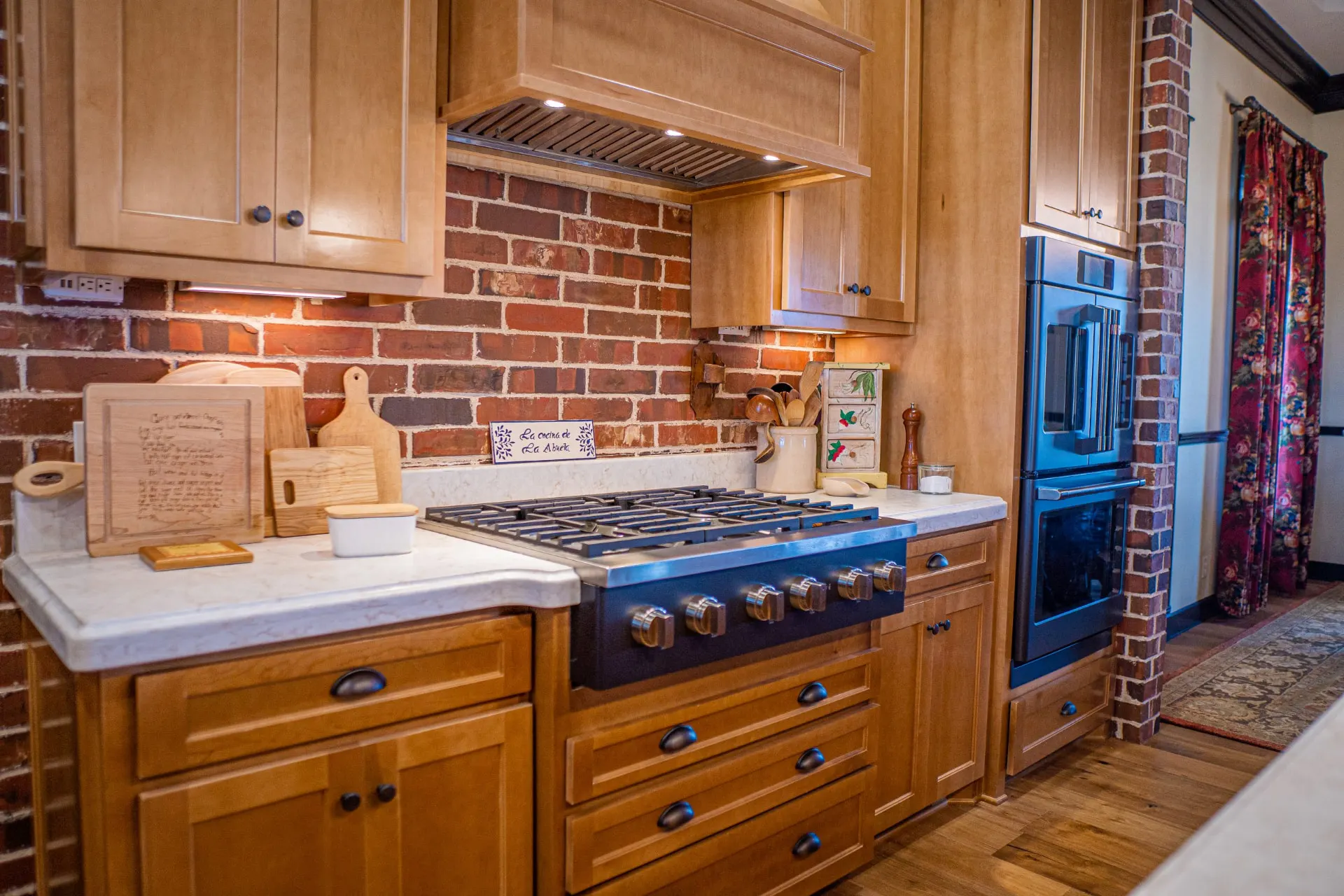 Kitchen cooking zone at the Wraparound Porch Home with a gas cooktop, a custom wood range hood and built-in double ovens against a red brick backsplash