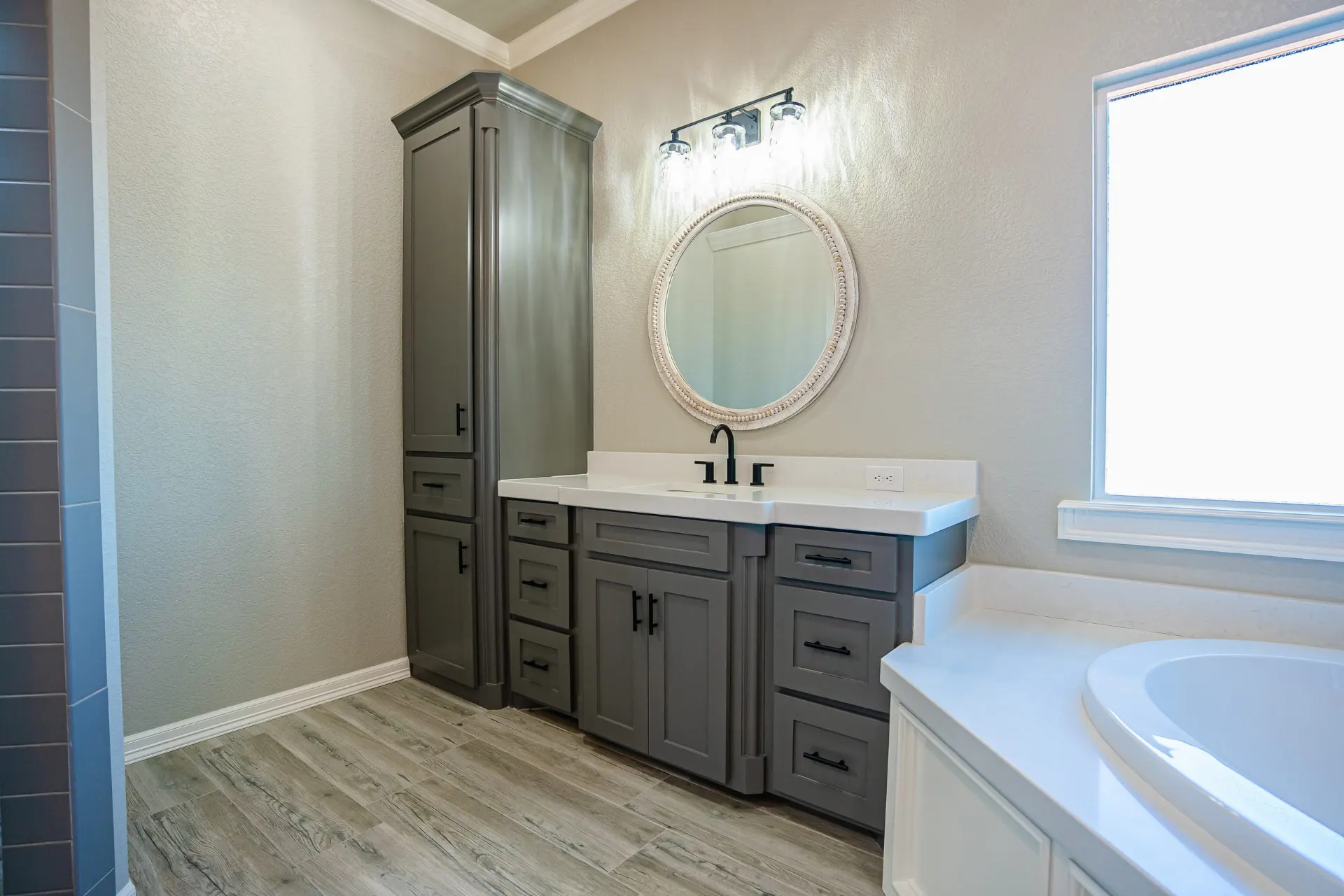 Primary bath vanity at the Eliam St Home with grey cabinetry, black hardware, a white-beaded round mirror and a tall linen cabinet