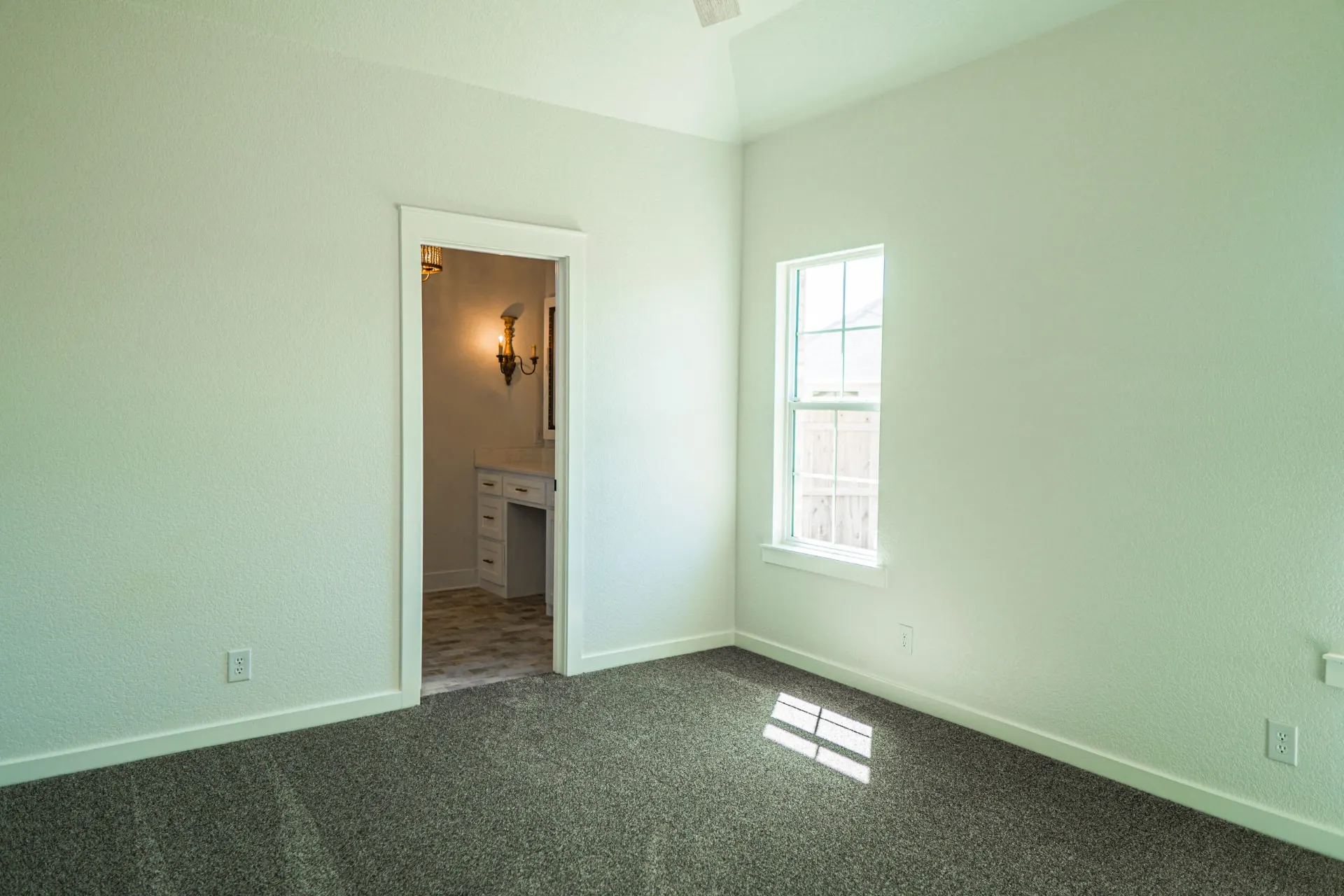 Empty carpeted bedroom at Sarai Home with a window view to the fenced yard and an open doorway to the ensuite vanity