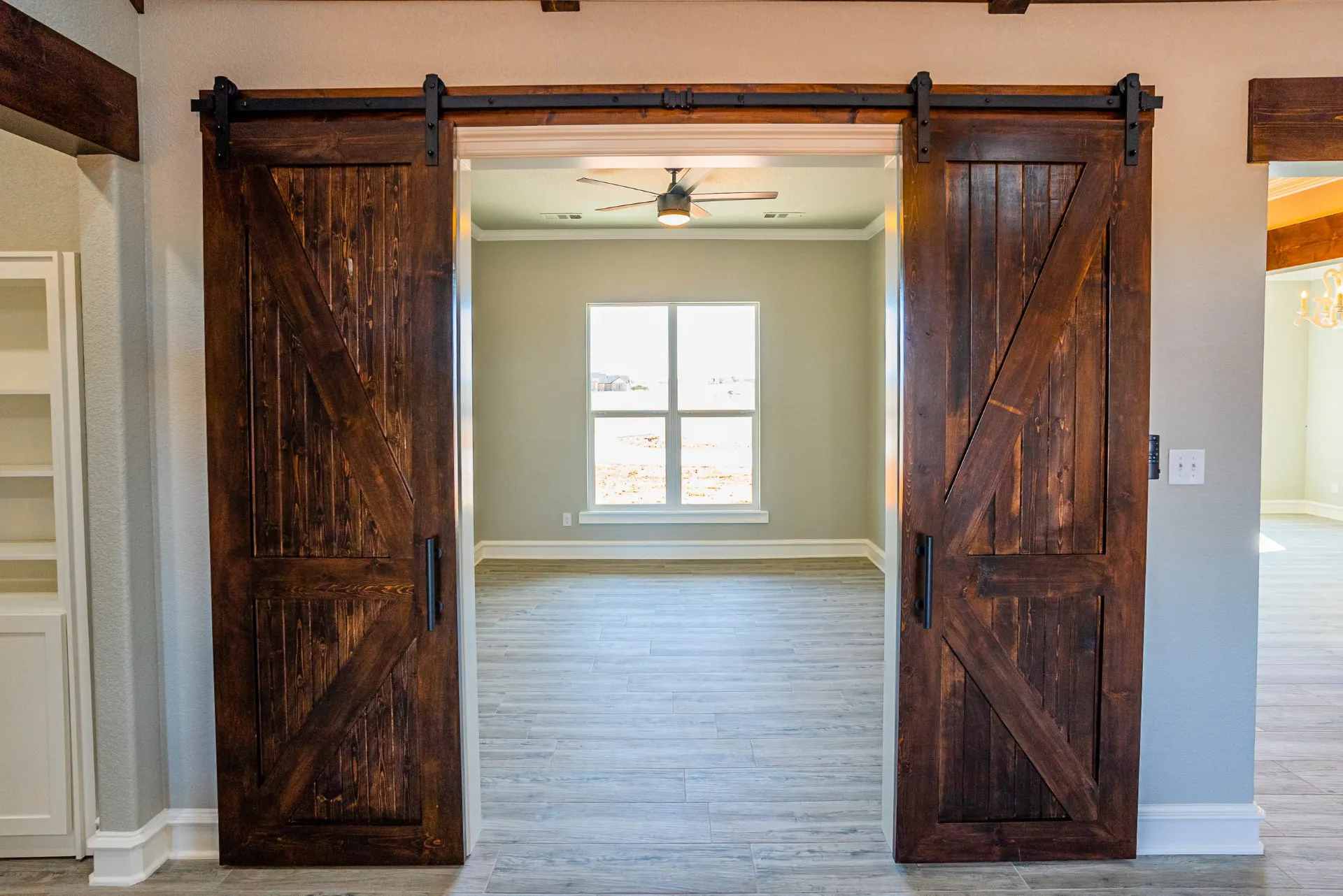 Double stained barn doors at the Eliam St Home opening into a flex room with grey walls, wood-look tile and a dark ceiling fan