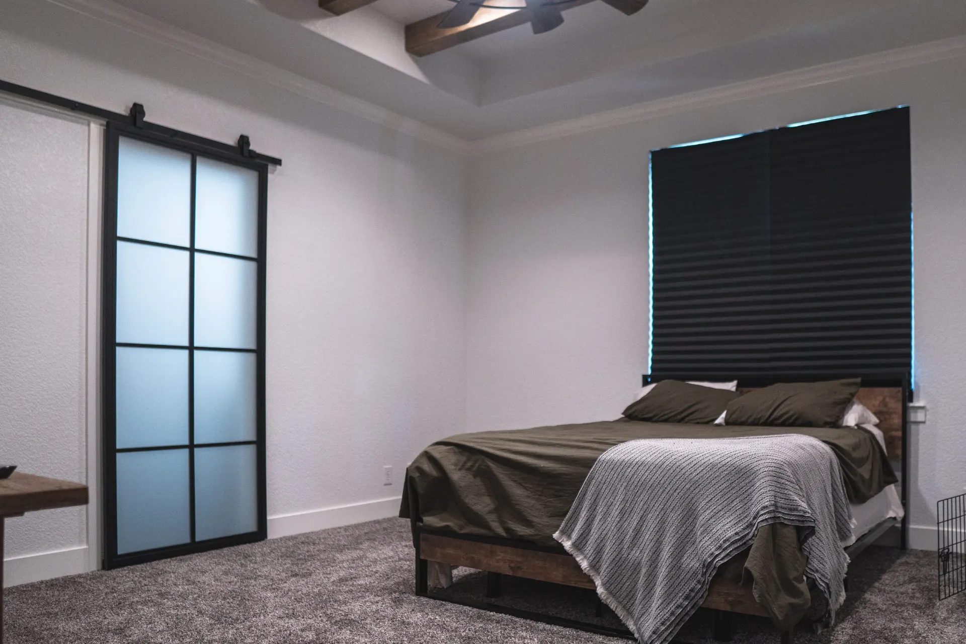 Bedroom at the K Ave Farmhouse with a black-frame glass barn door, olive bedding on a wood bed frame, black cellular shades and gray carpet