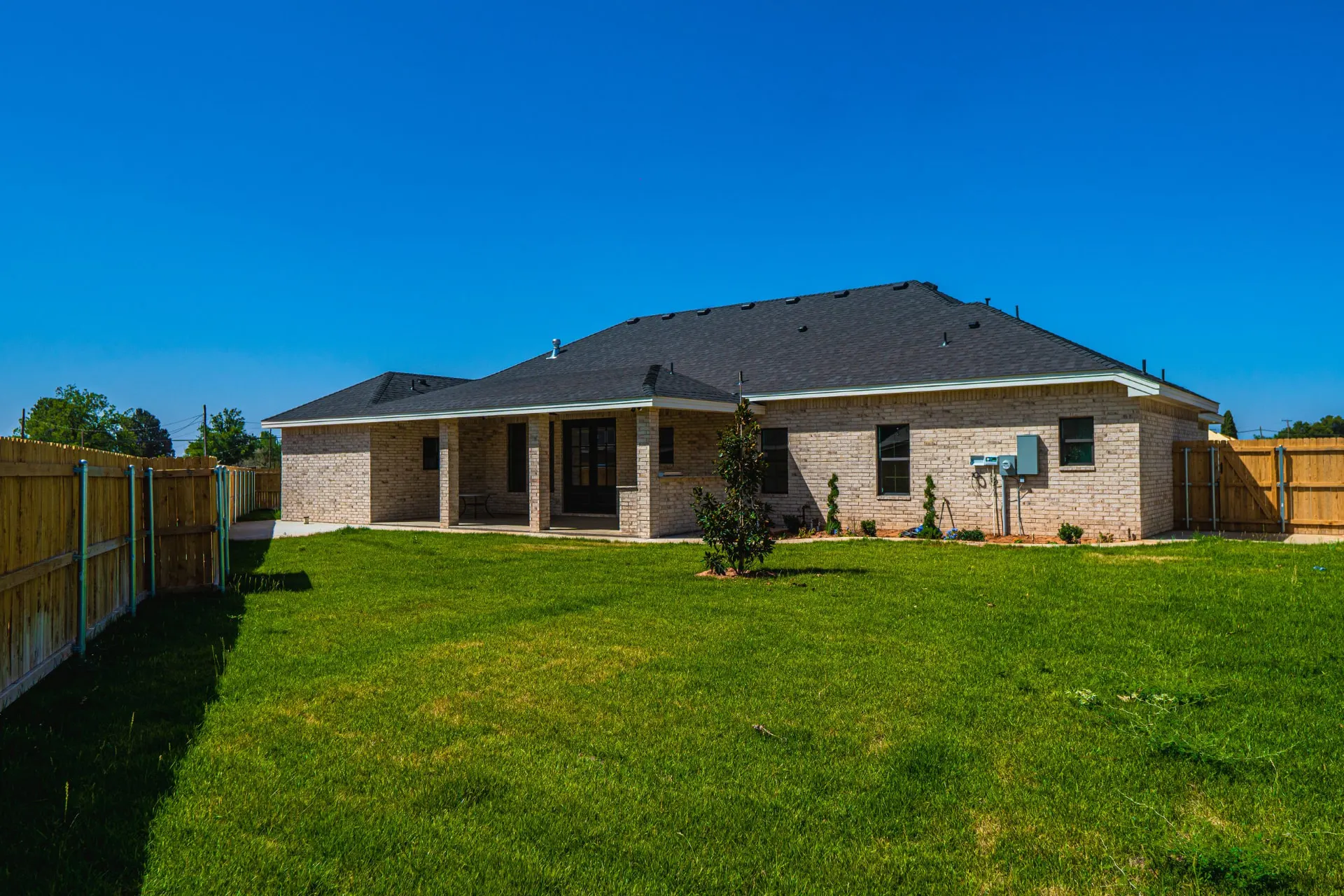 Rear elevation of Sarai Home in light brick with a covered patio under the hip roof, a young magnolia and a fenced back lawn