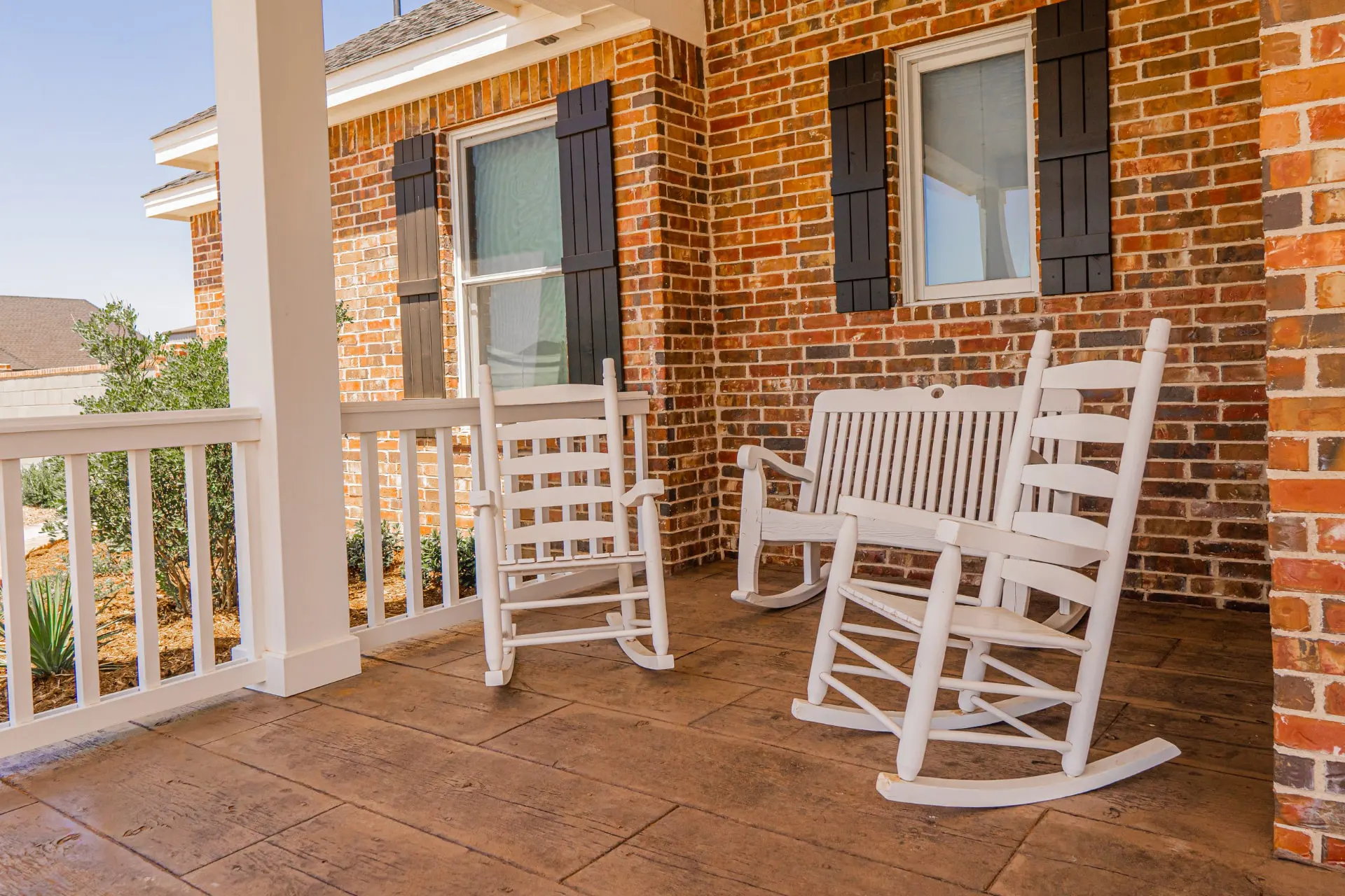 Front porch at the Wraparound Porch Home with white rocking chairs and a porch bench on stamped concrete against red brick and black shutters