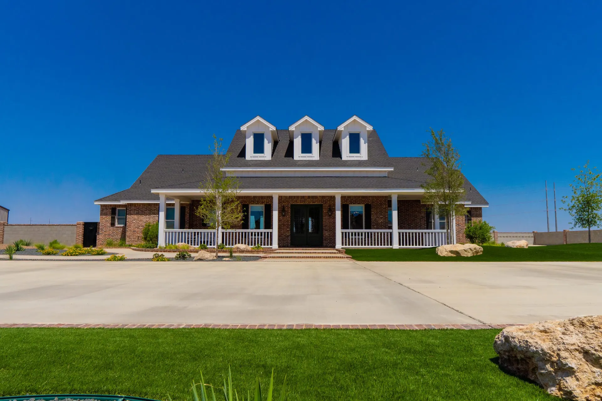 Front elevation of the Wraparound Porch Home with white porch columns, three gabled dormers and a wide concrete drive flanked by fresh landscaping