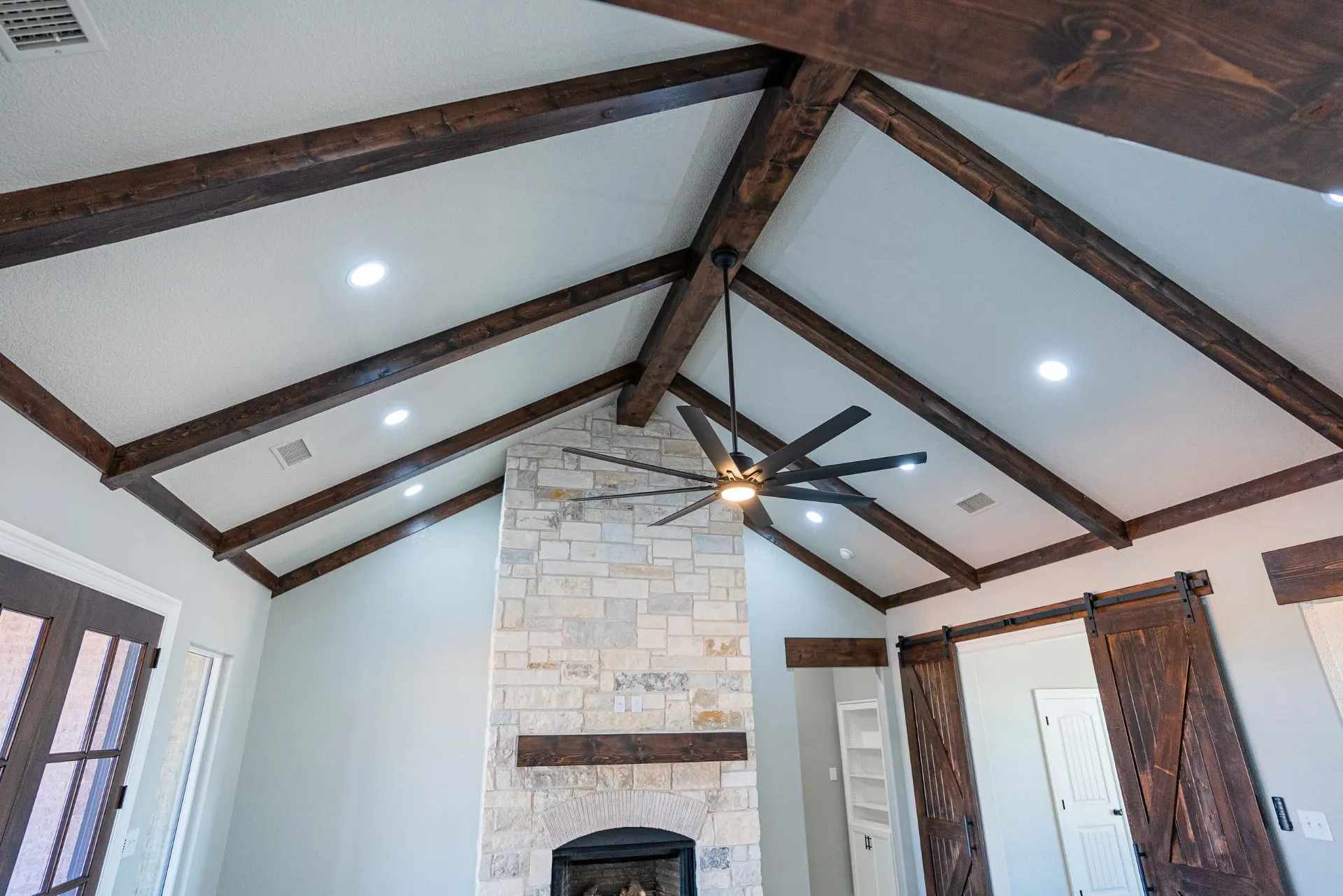 Vaulted great room ceiling at the Eliam St Home with dark-stained wood trusses, recessed lights and a black fan above the full-height stone fireplace and barn doors