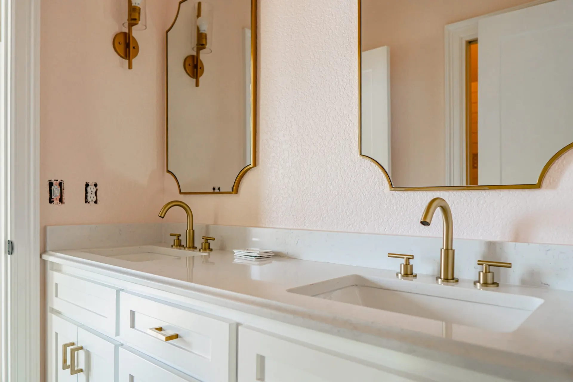 Double vanity bath at the 12th St Home with white cabinets, brushed gold faucets and scalloped mirrors on blush textured walls
