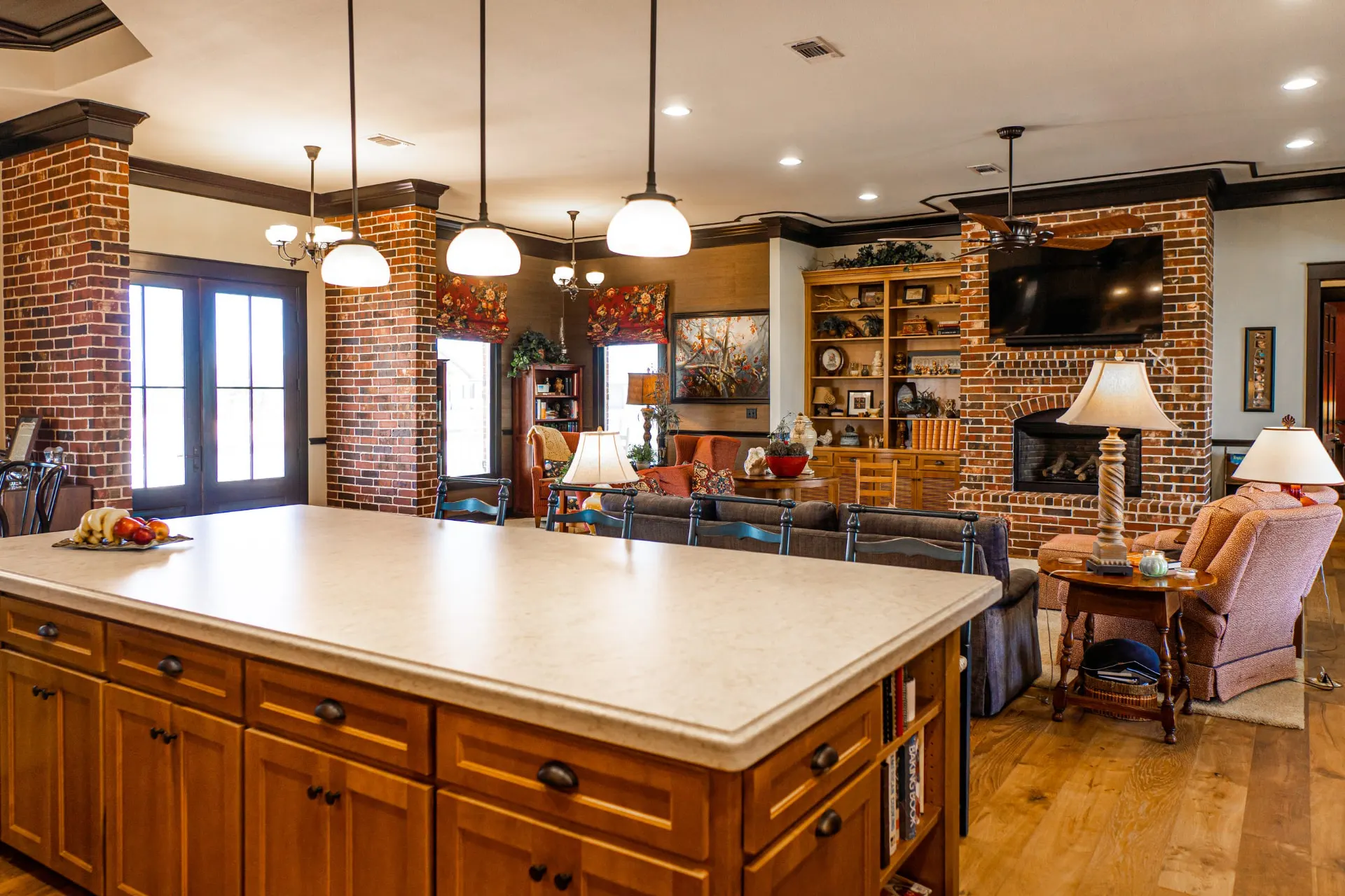 View across the kitchen island at the Wraparound Porch Home toward the living room, with a brick fireplace, wall-mounted television and built-in bookshelves