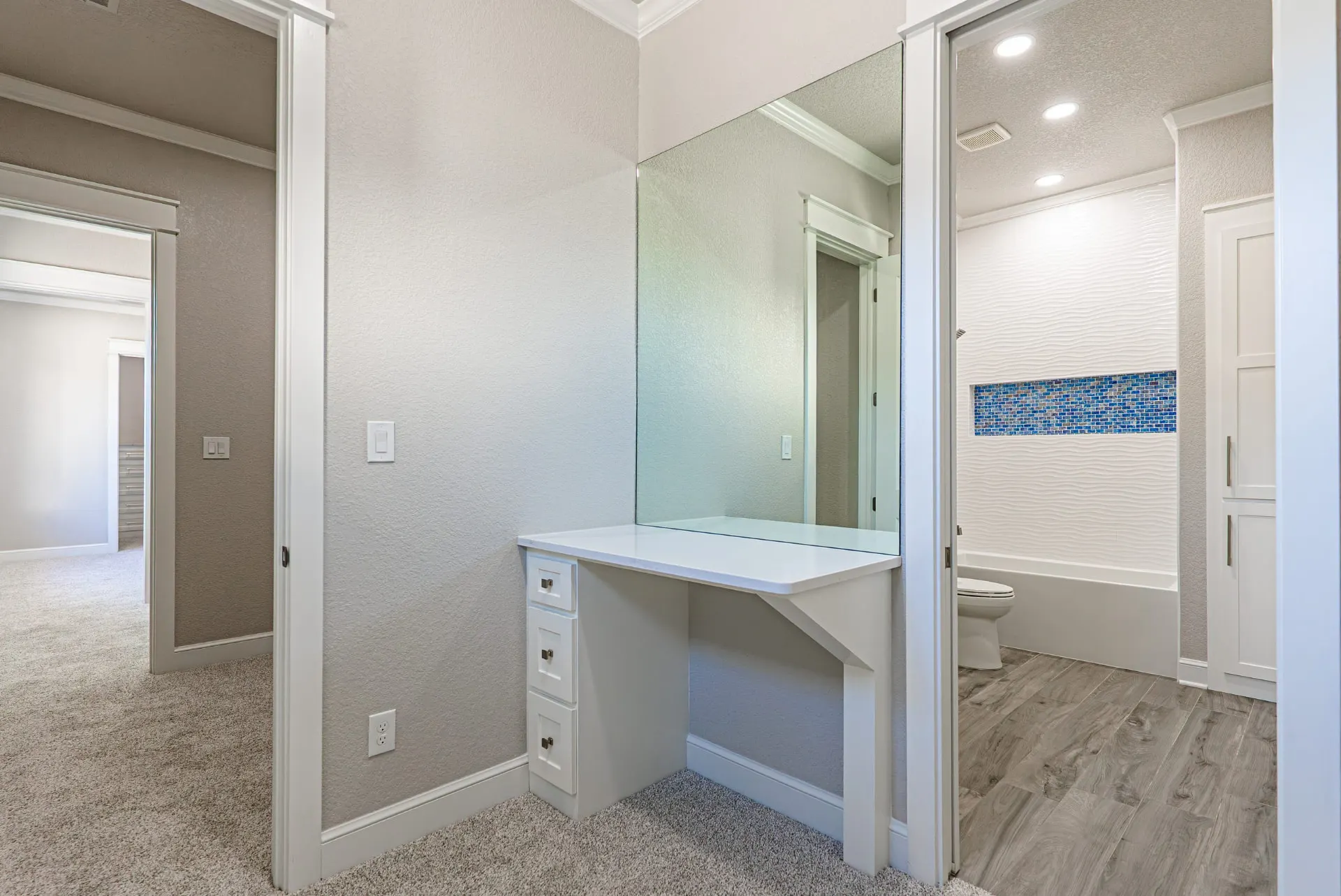 Dressing nook at 27 Santiago Mission Dr with a built-in white vanity and frameless mirror, opening to a bath with wavy tile and a blue mosaic niche