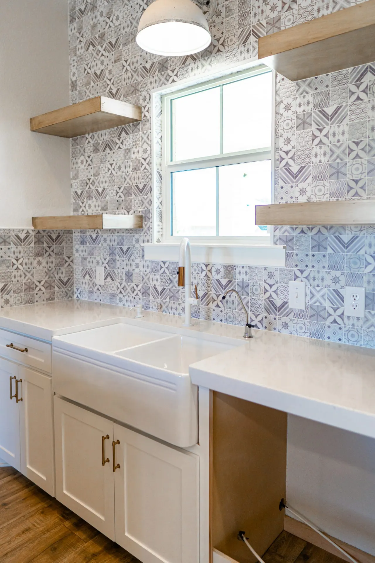 Farmhouse sink at Sarai Home set under the window in the patterned-tile backsplash wall, with wood floating shelves and a polished chrome drinking faucet