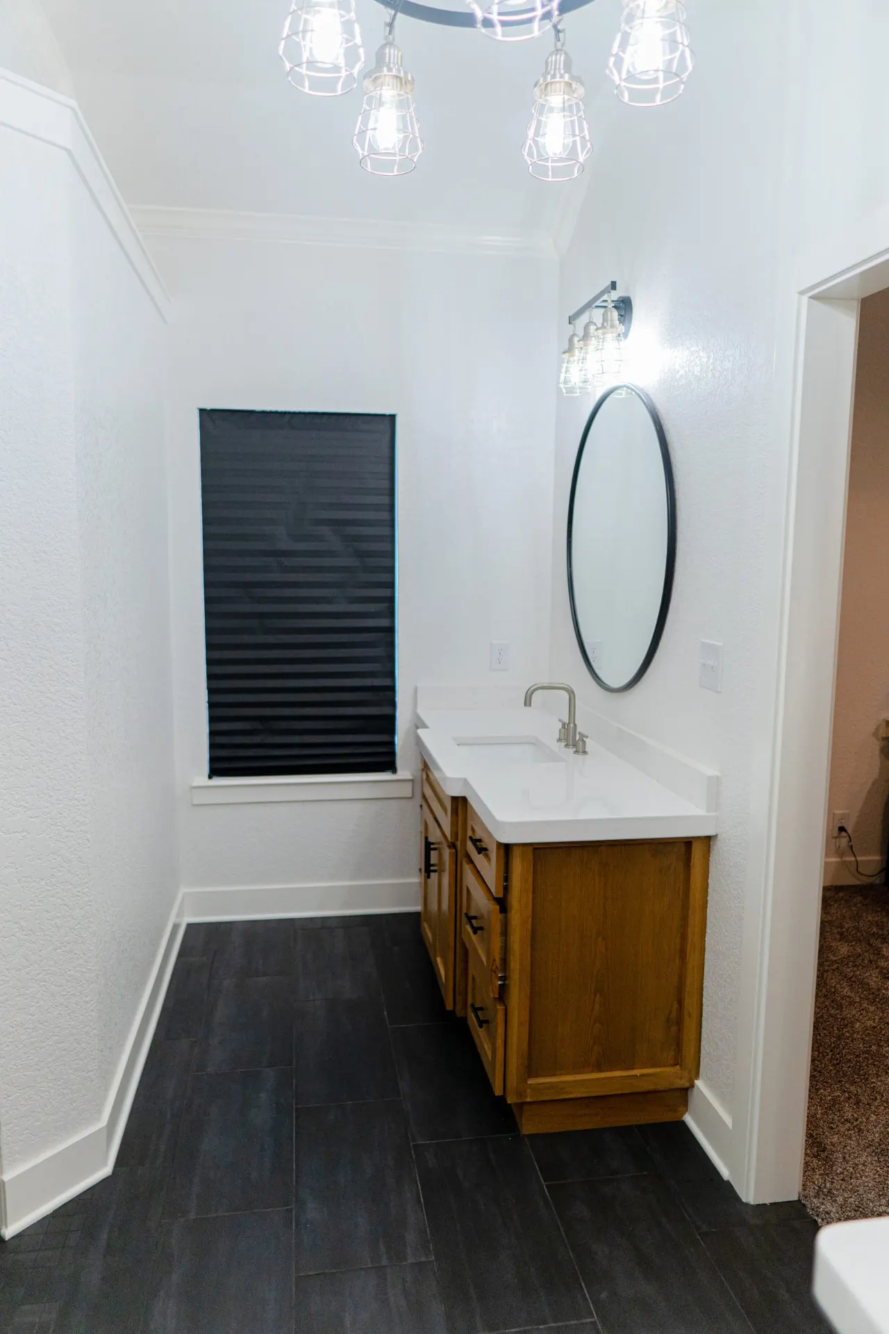 Bathroom at the K Ave Farmhouse with a honey-oak vanity, white quartz top, black-framed oval mirror, wire-cage chandelier and dark wood-look floor tile