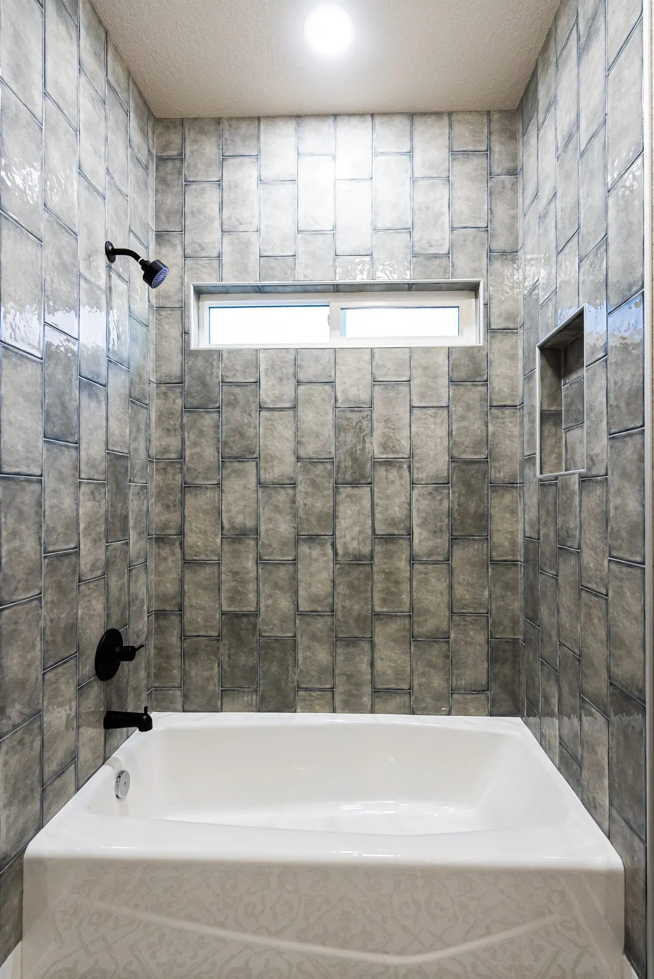 Tub at 708 K Ave wrapped in gray handcrafted tile with black fixtures, a niche shelf and a transom window above