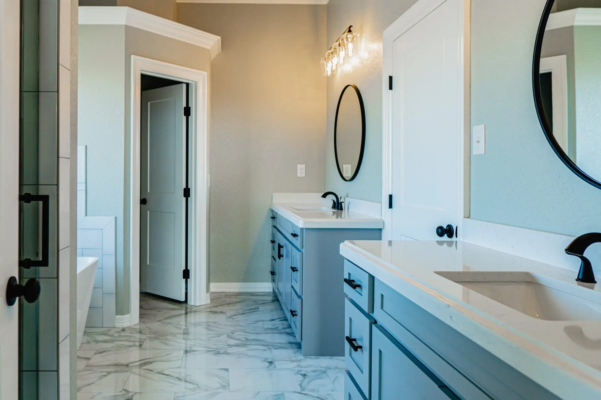 Primary bath at the 12th St Home with grey double vanities, black fixtures, round mirrors, marble-look tile and a freestanding tub