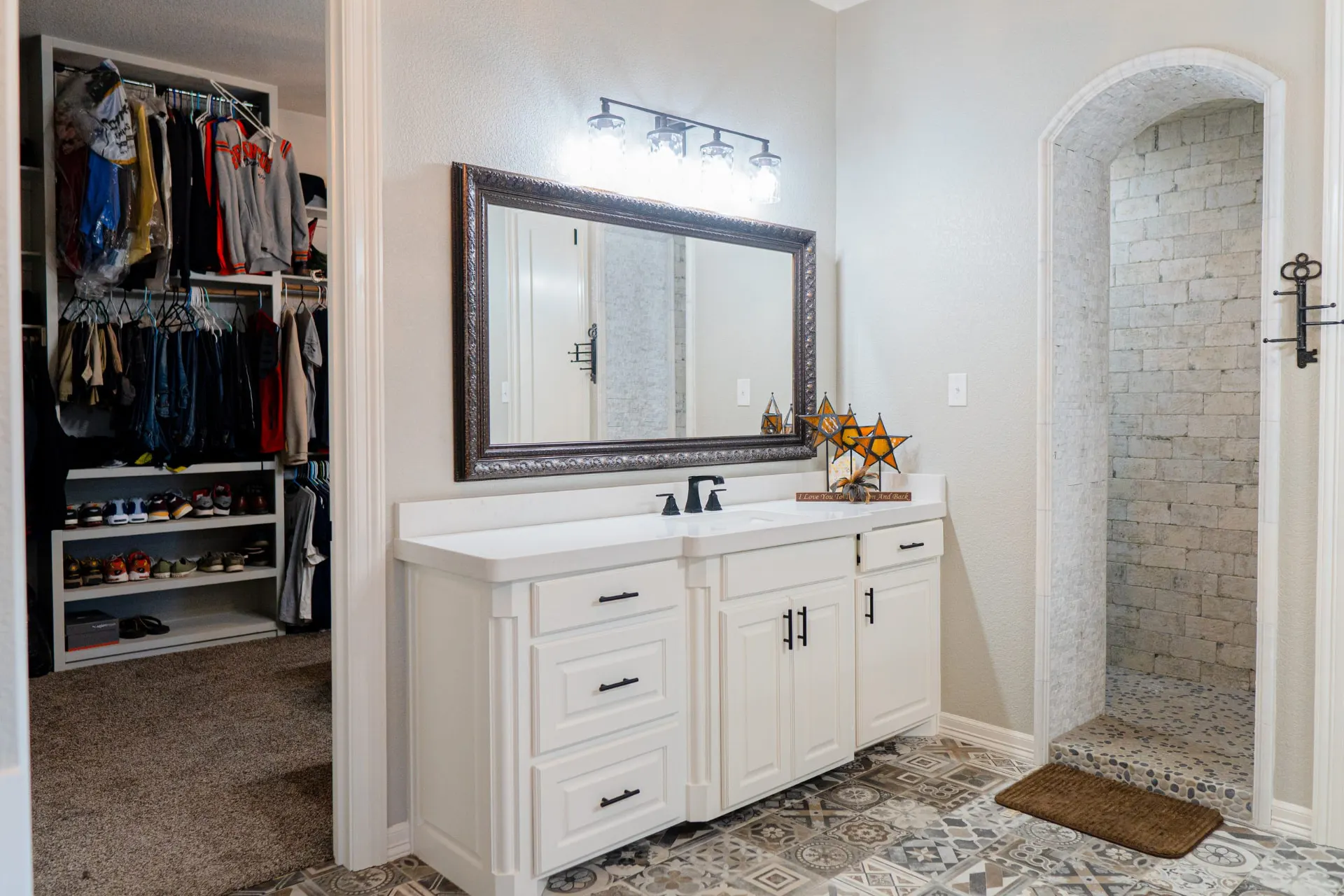Primary vanity in white raised-panel cabinetry with a bronze ornate mirror, beside an arched stone-tile walk-in shower and the walk-in closet at 8821 Shawn Dr