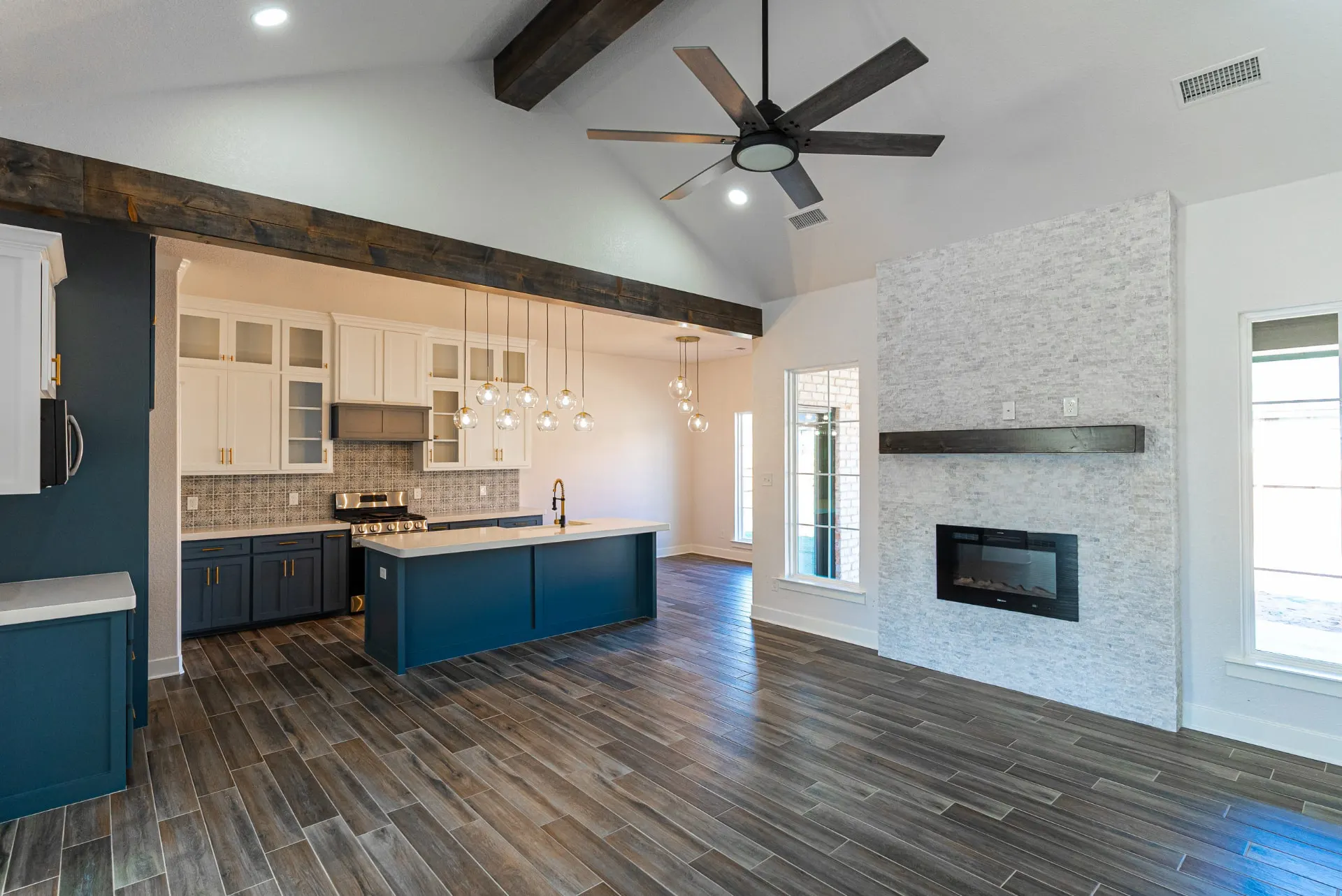 Open great room and kitchen at 718 K Ave under a vaulted ceiling with a rough-hewn beam, teal island, glass globe pendants and a stacked-stone fireplace