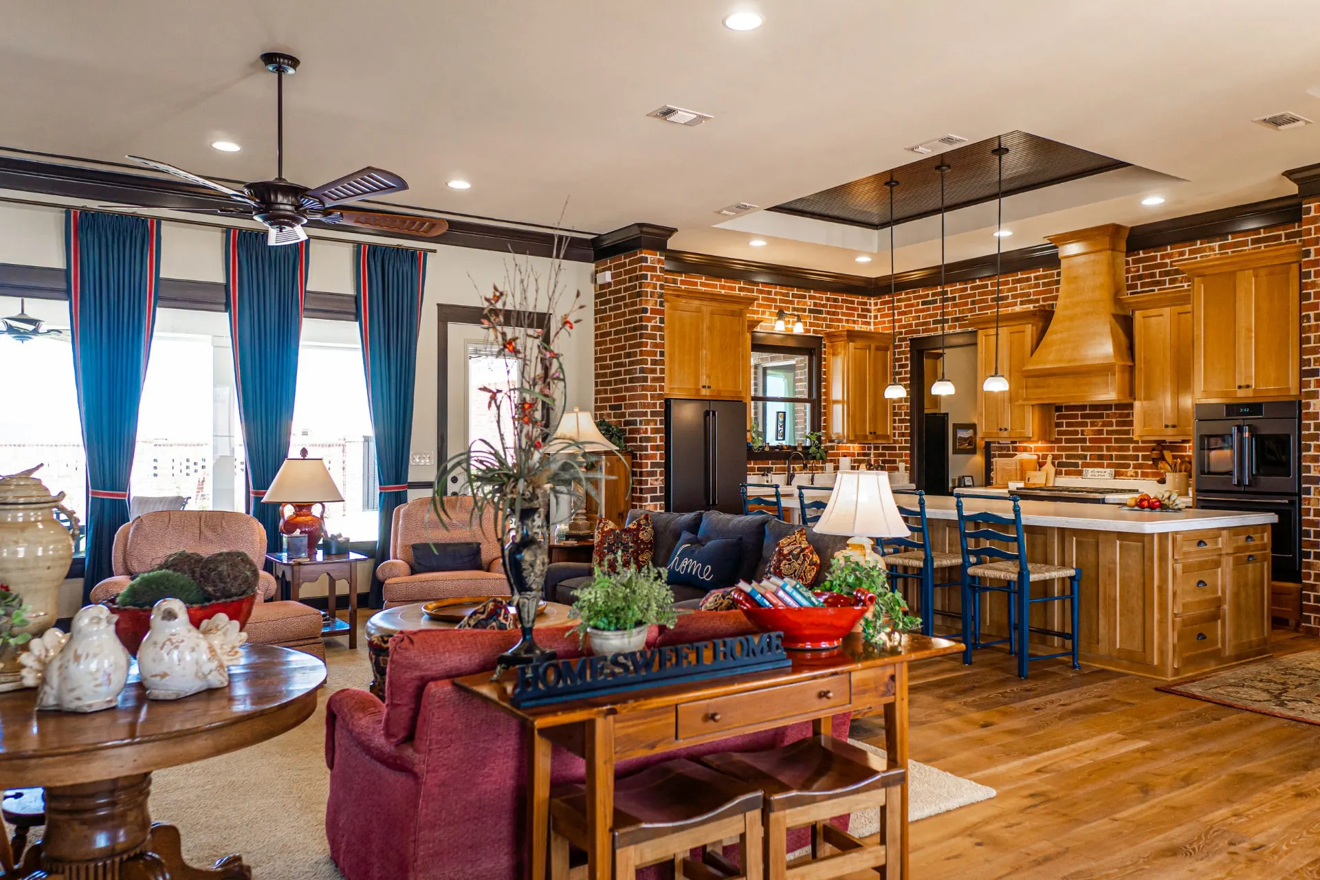 Open living room at the Wraparound Porch Home with navy and cranberry seating, a brick fireplace column and the kitchen island with glass pendants beyond