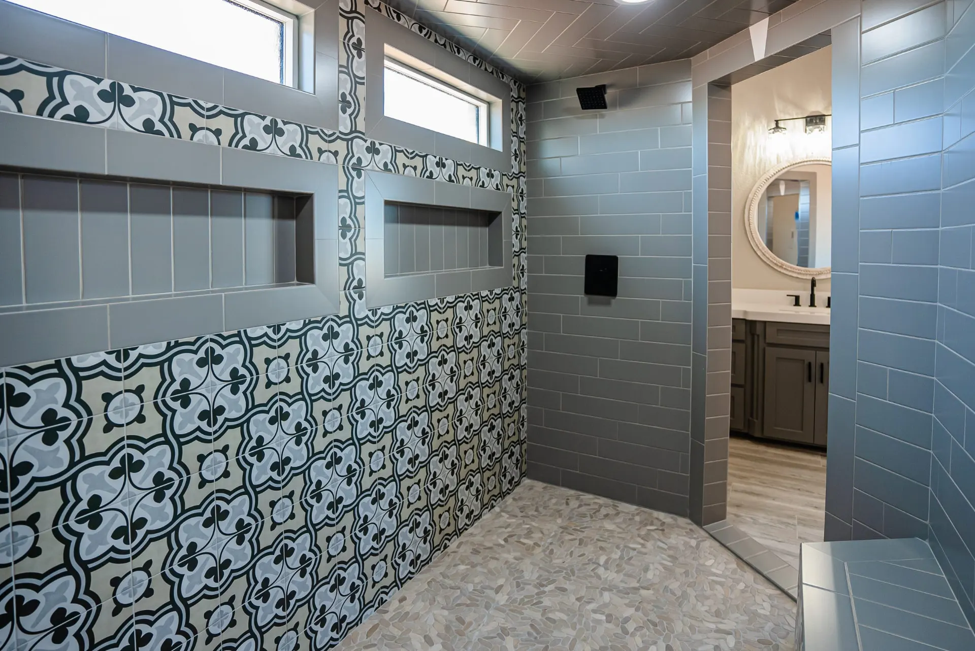 Wide view of the Eliam St Home walk-in shower with quatrefoil tile, grey subway walls, pebble flooring and the vanity beyond