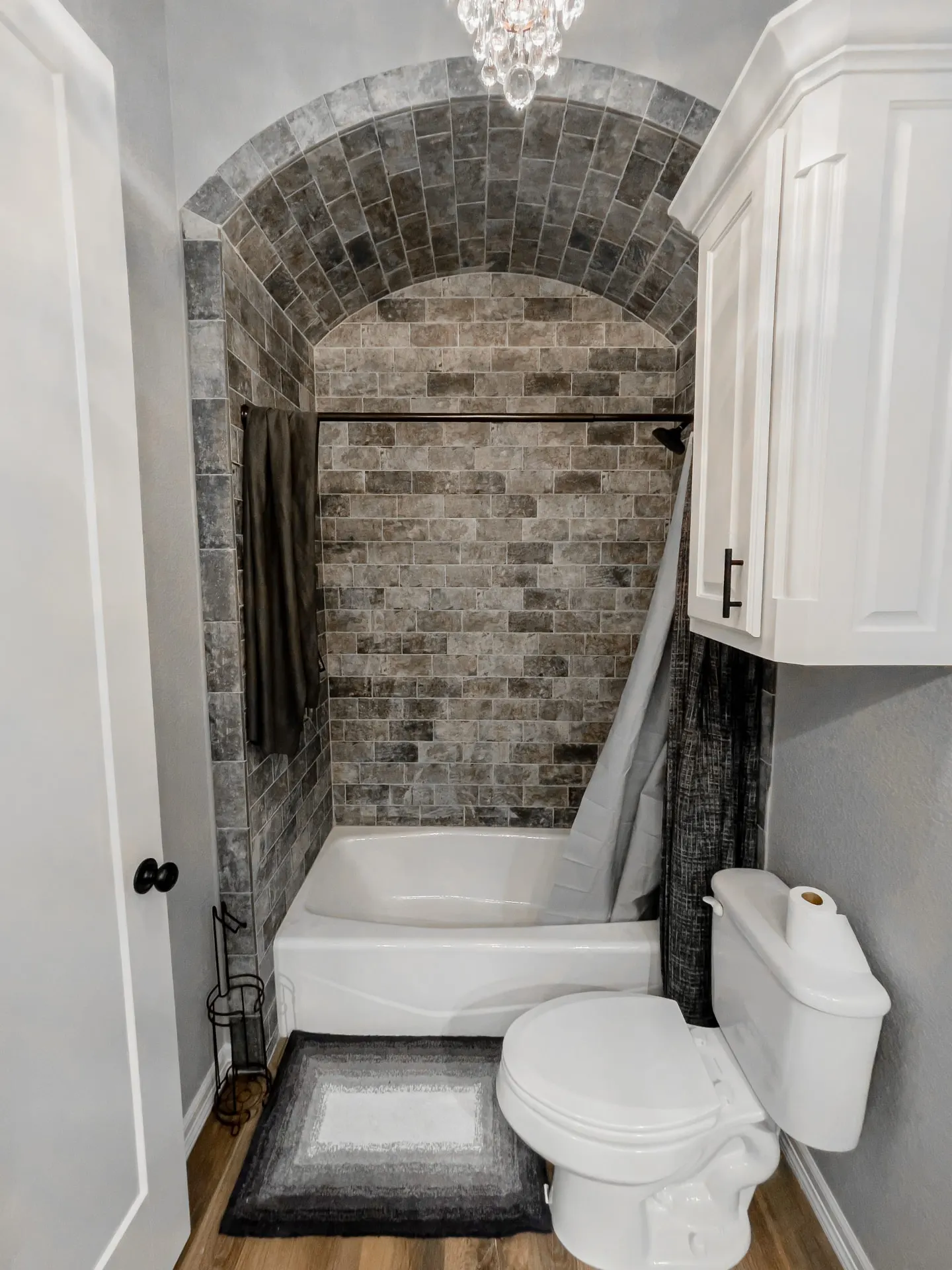 Tub and shower at 8821 Shawn Dr under a barrel-vaulted alcove lined in rustic brick-look tile, with a mini crystal chandelier at the peak